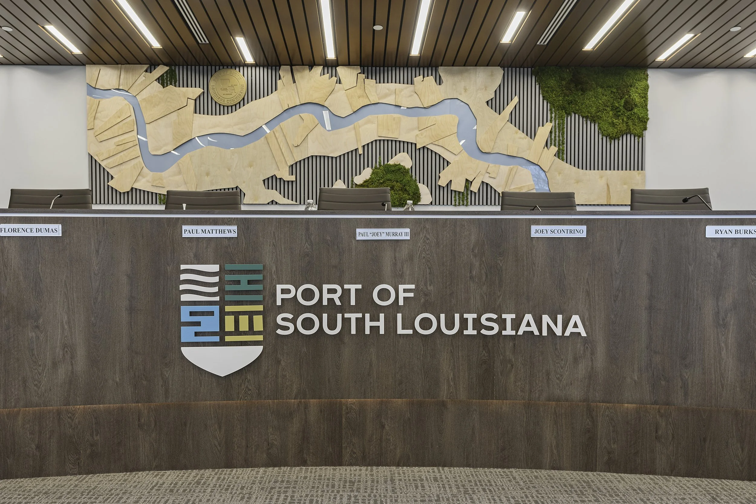 Wooden map of the Port of South Louisiana with water channels and greenery, mounted on a wall behind a panel table with signs for officials.