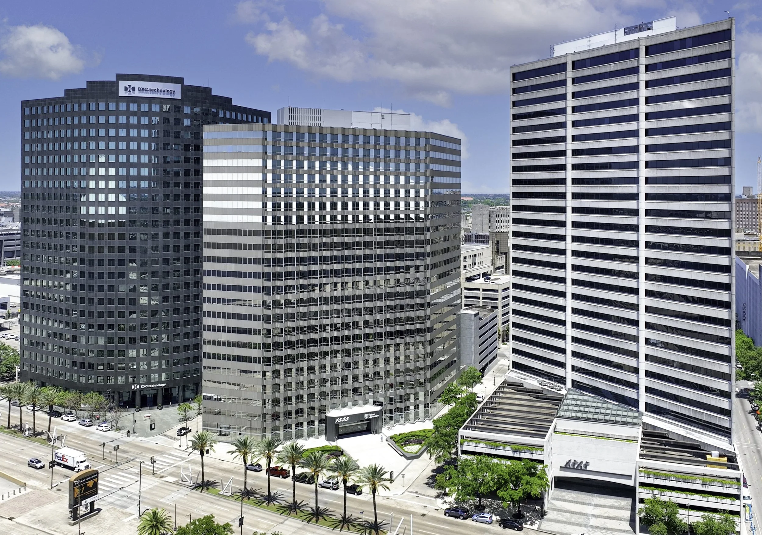 Cityscape with tall office buildings, a busy street with cars and palm trees, under a partly cloudy sky.