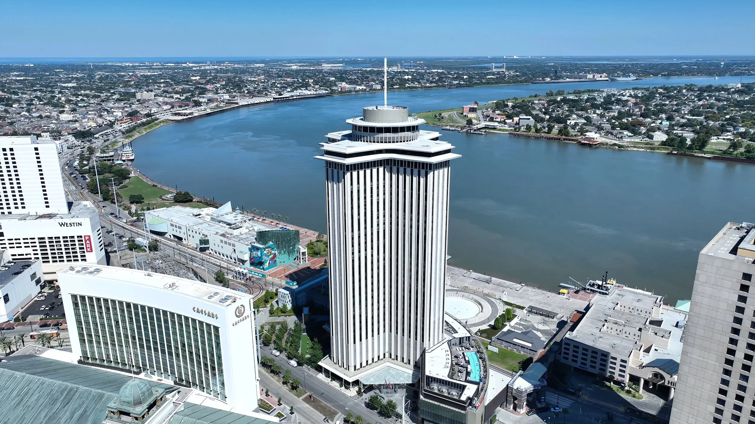 Aerial view of a city skyline featuring a tall building with horizontal lines, a river, and a green park area along the waterfront.