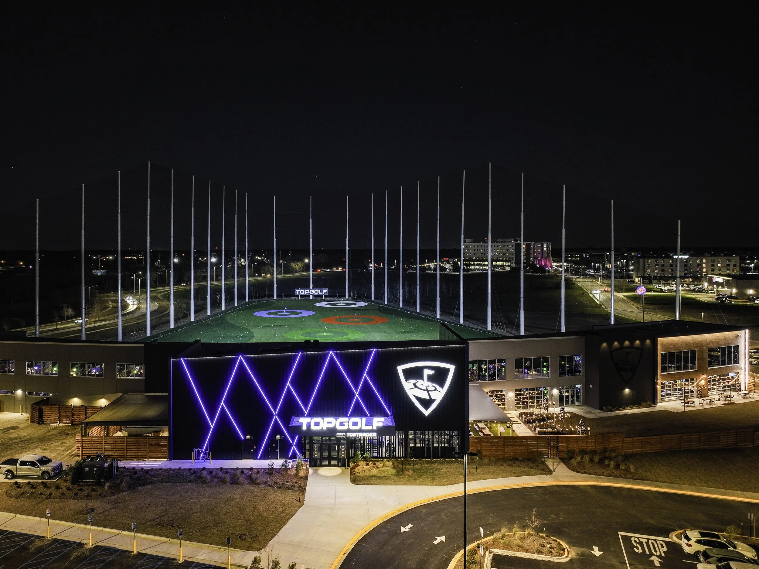 Night view of Topgolf entertainment complex with illuminated signage, modern architecture, and a golf practice field, with city lights in the background.