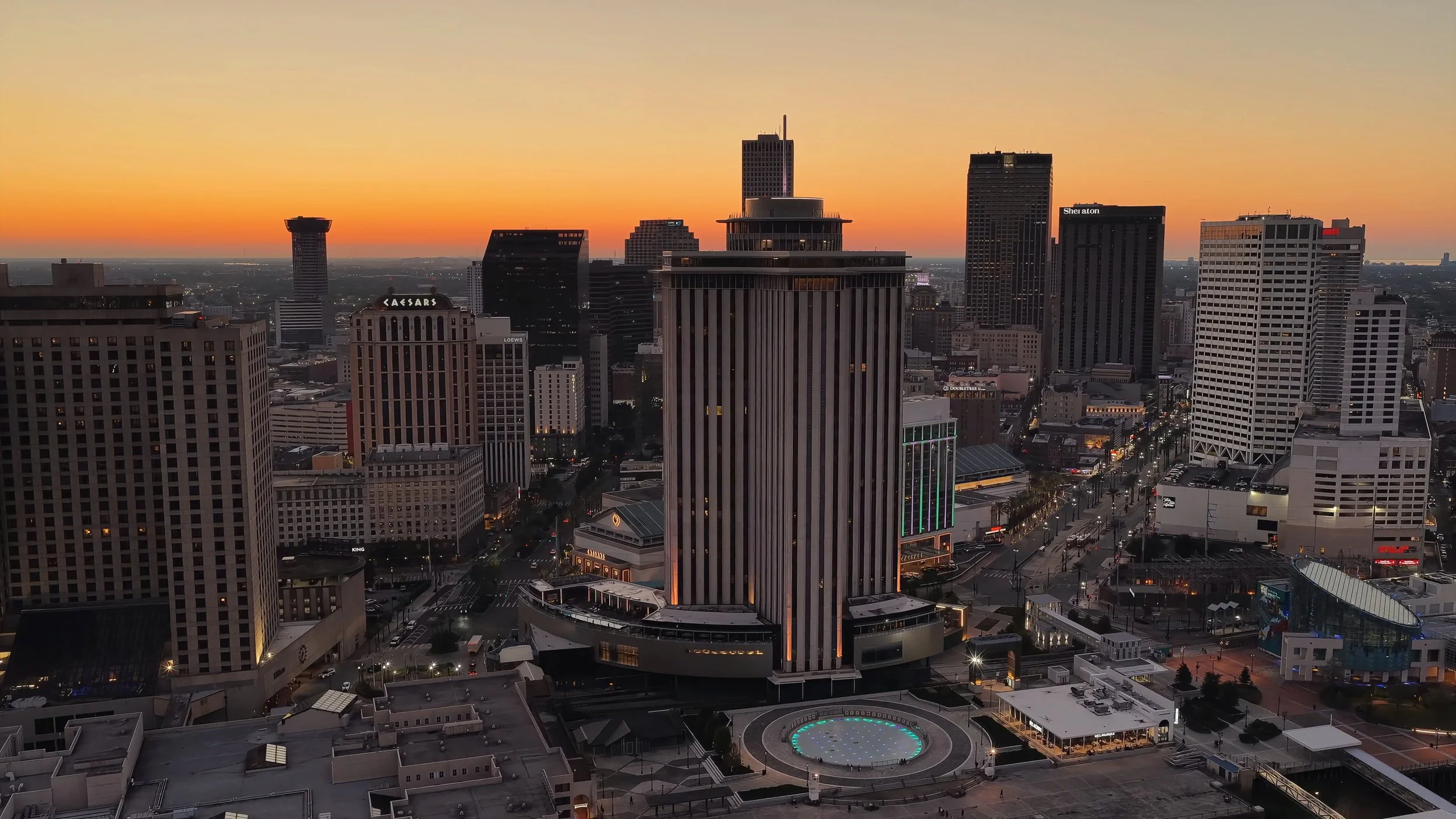 View of a city skyline at sunset with tall buildings, including the Caesars and Sheraton hotels, and a lit-up circular structure in the foreground.