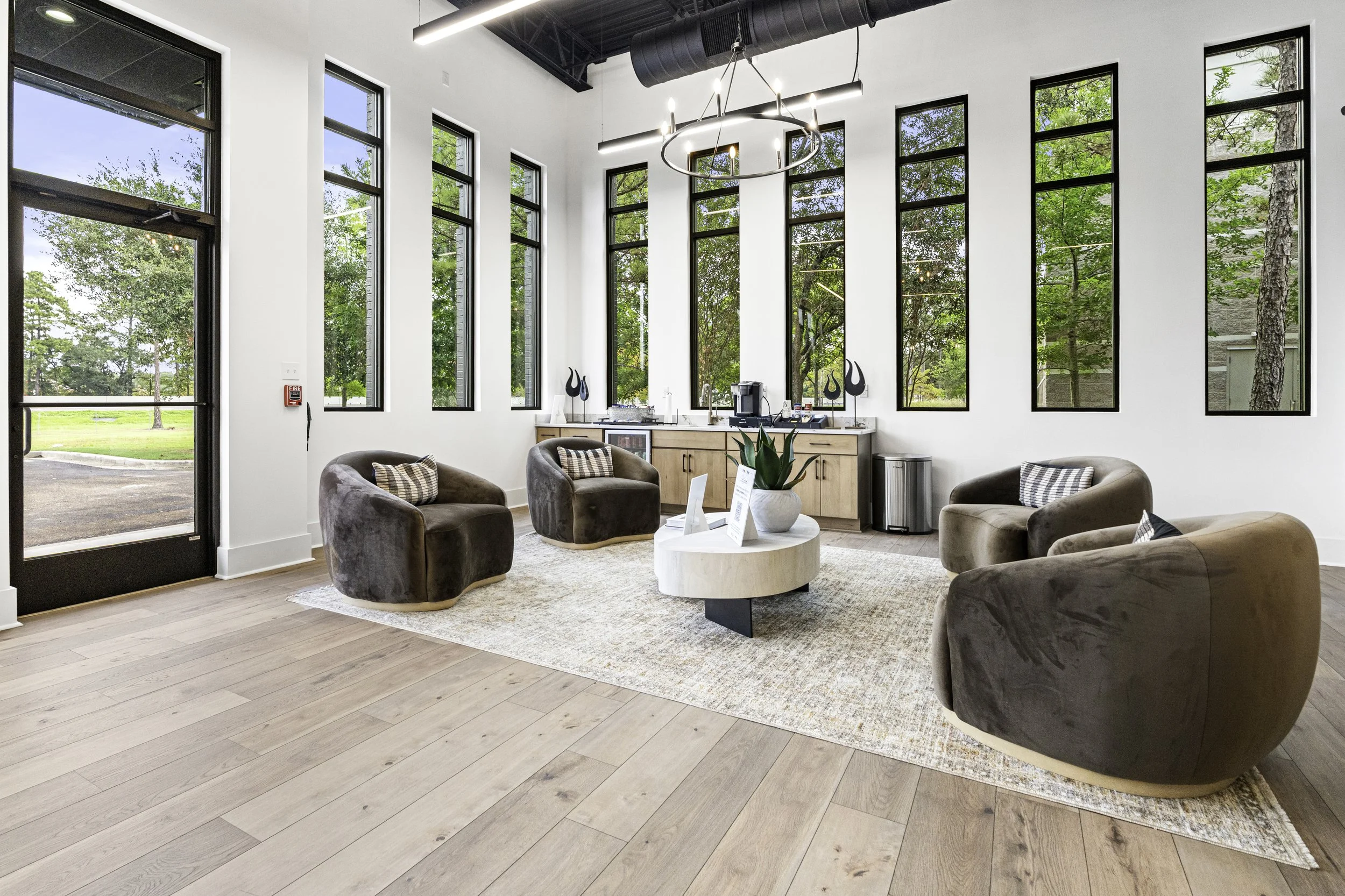 Modern lounge with multiple large tall windows, neutral-colored couches, a round coffee table, and a kitchenette area in the back, with a view of trees outside.
