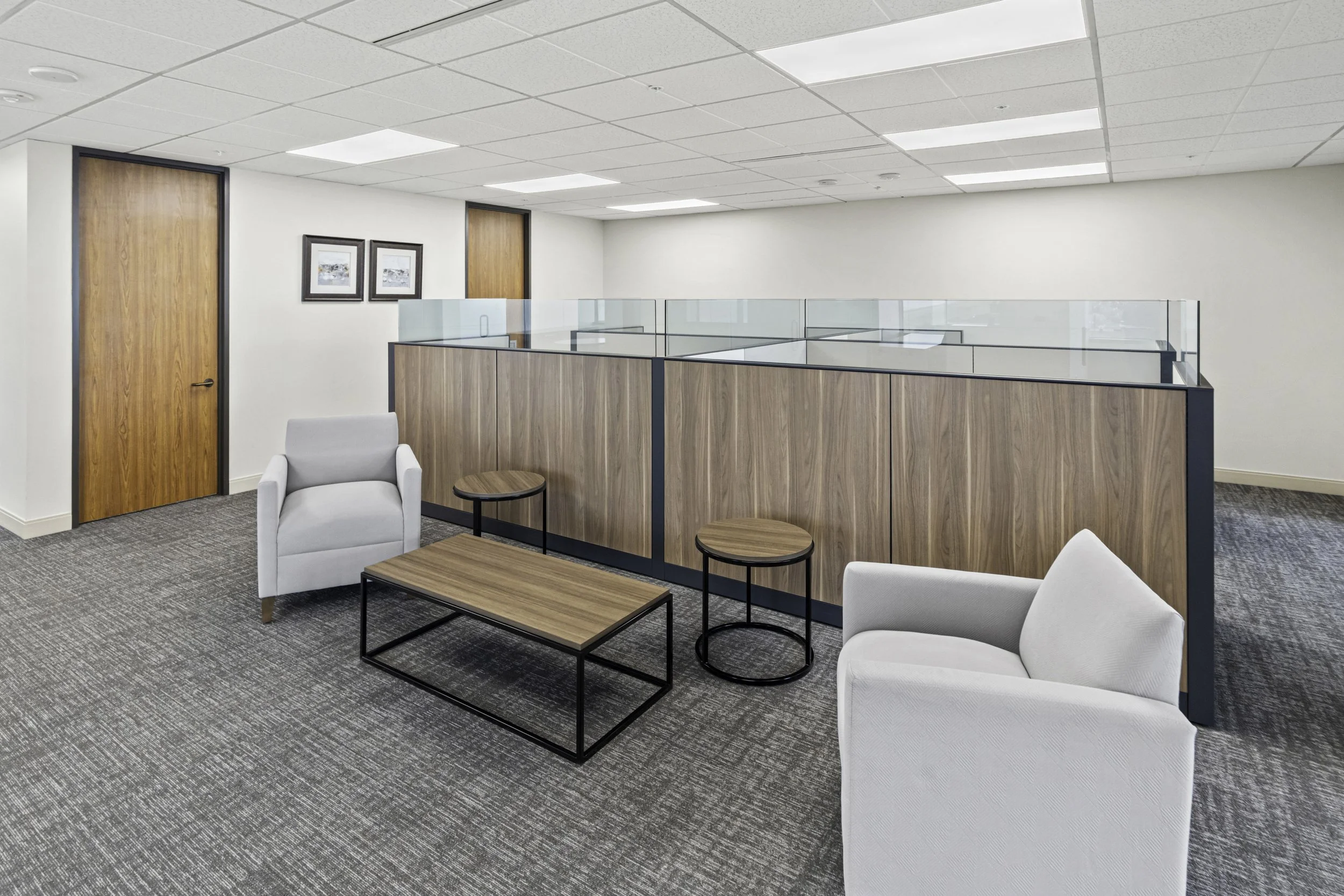 Empty office space with white chairs, wooden tables, a glass partition, framed pictures on the wall, closed wooden doors, and grid ceiling with fluorescent lighting.