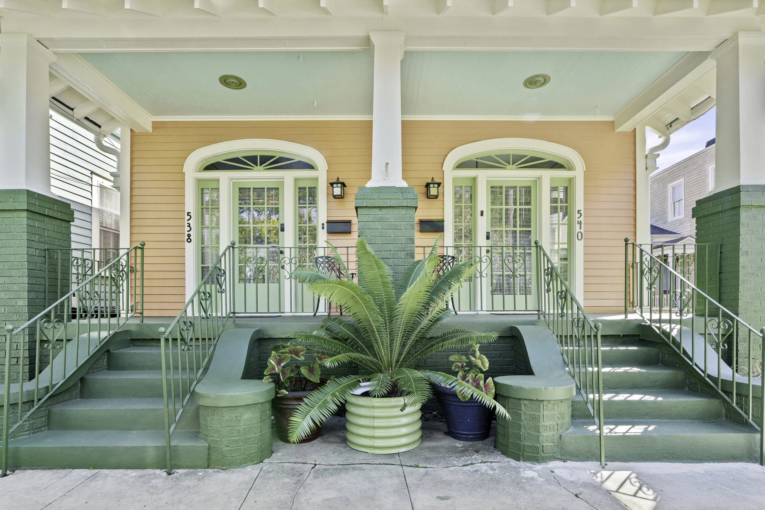 Front view of a pastel-colored house with two front doors, surrounded by potted plants and decorative green trim.