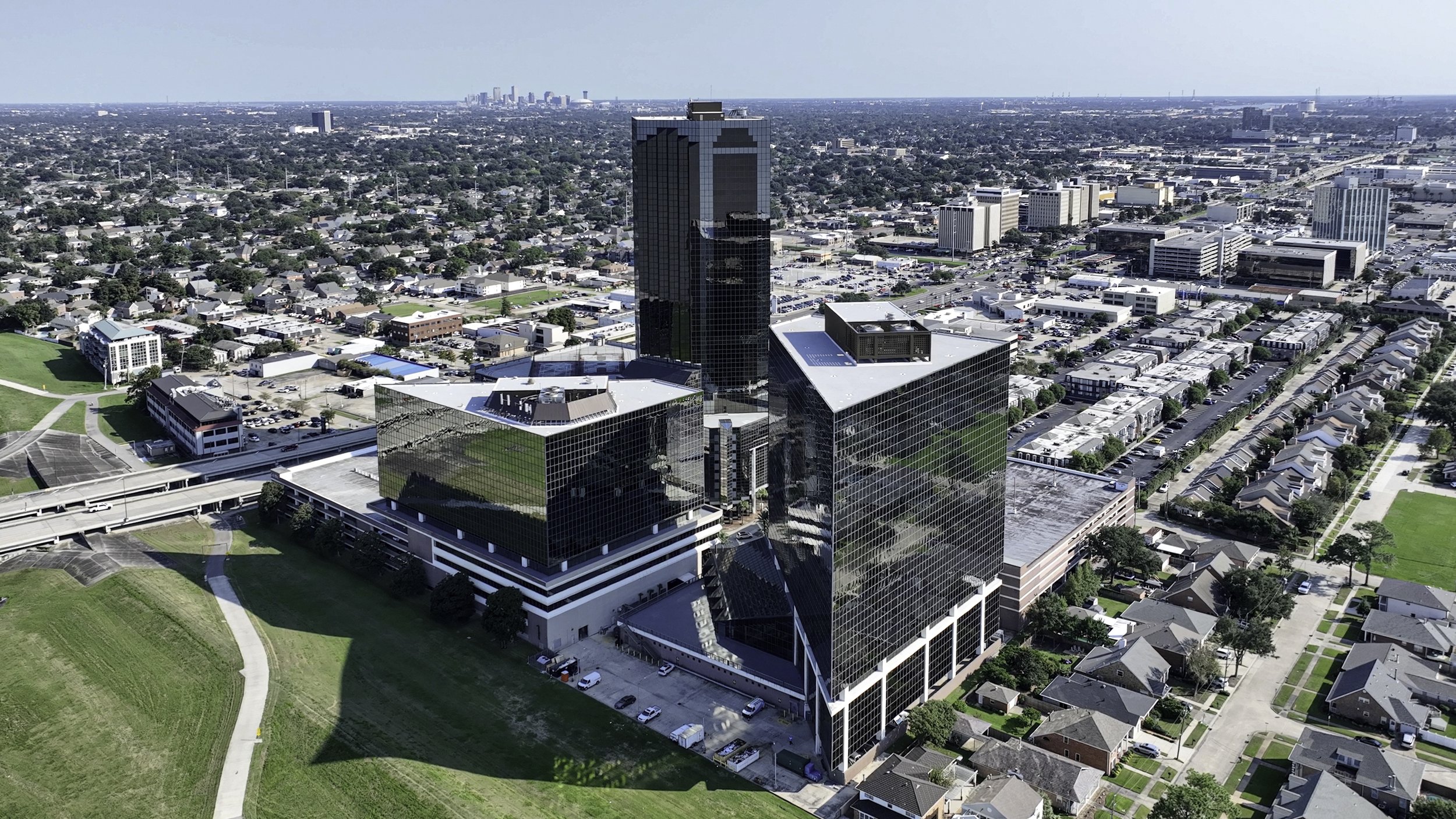 A cityscape featuring tall modern office buildings with glass facades, a high-rise in the center, and surrounding residential neighborhoods with houses and streets, under a clear blue sky.