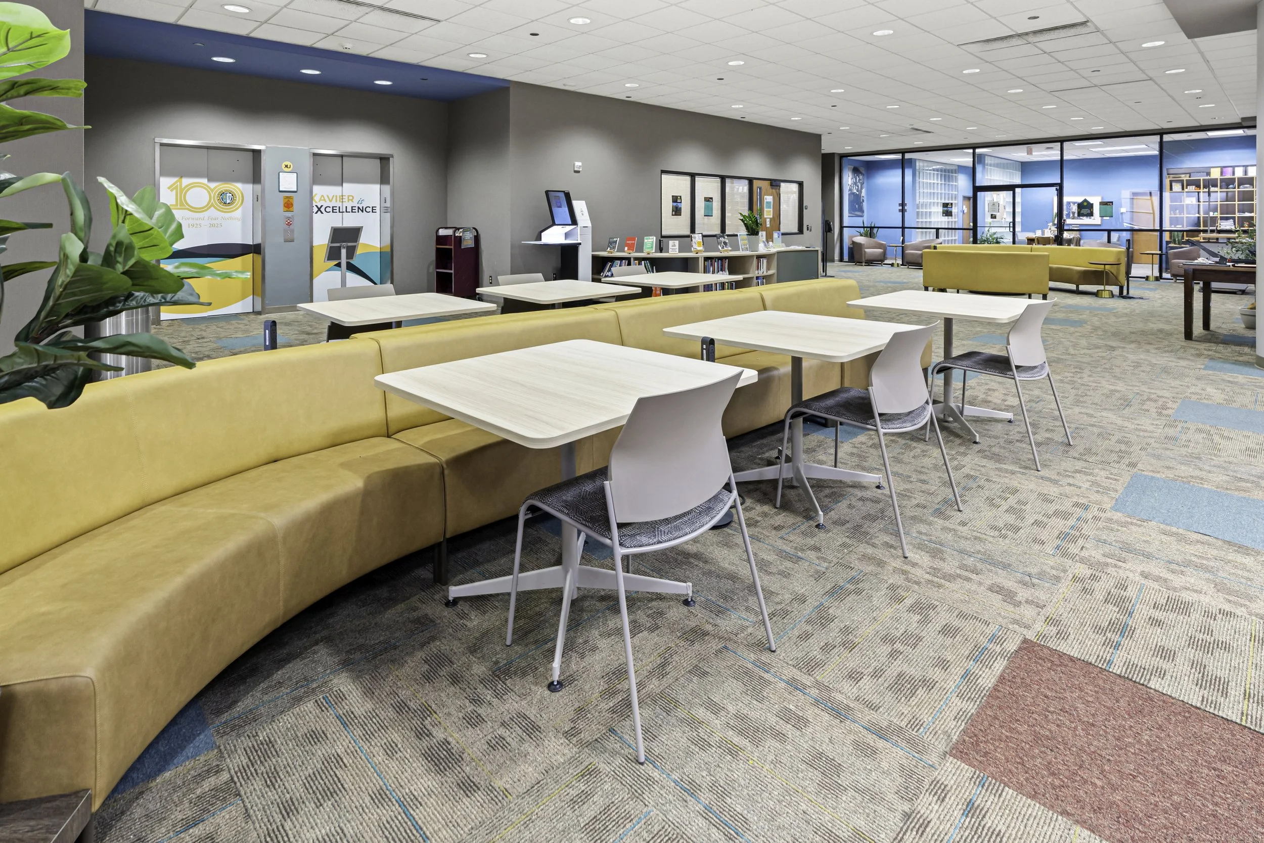 A spacious waiting area with yellow cushioned benches, white tables, and modern chairs. There is a bookshelf with books, digital kiosks, and large windows and glass doors leading to another room with blue walls and additional seating.