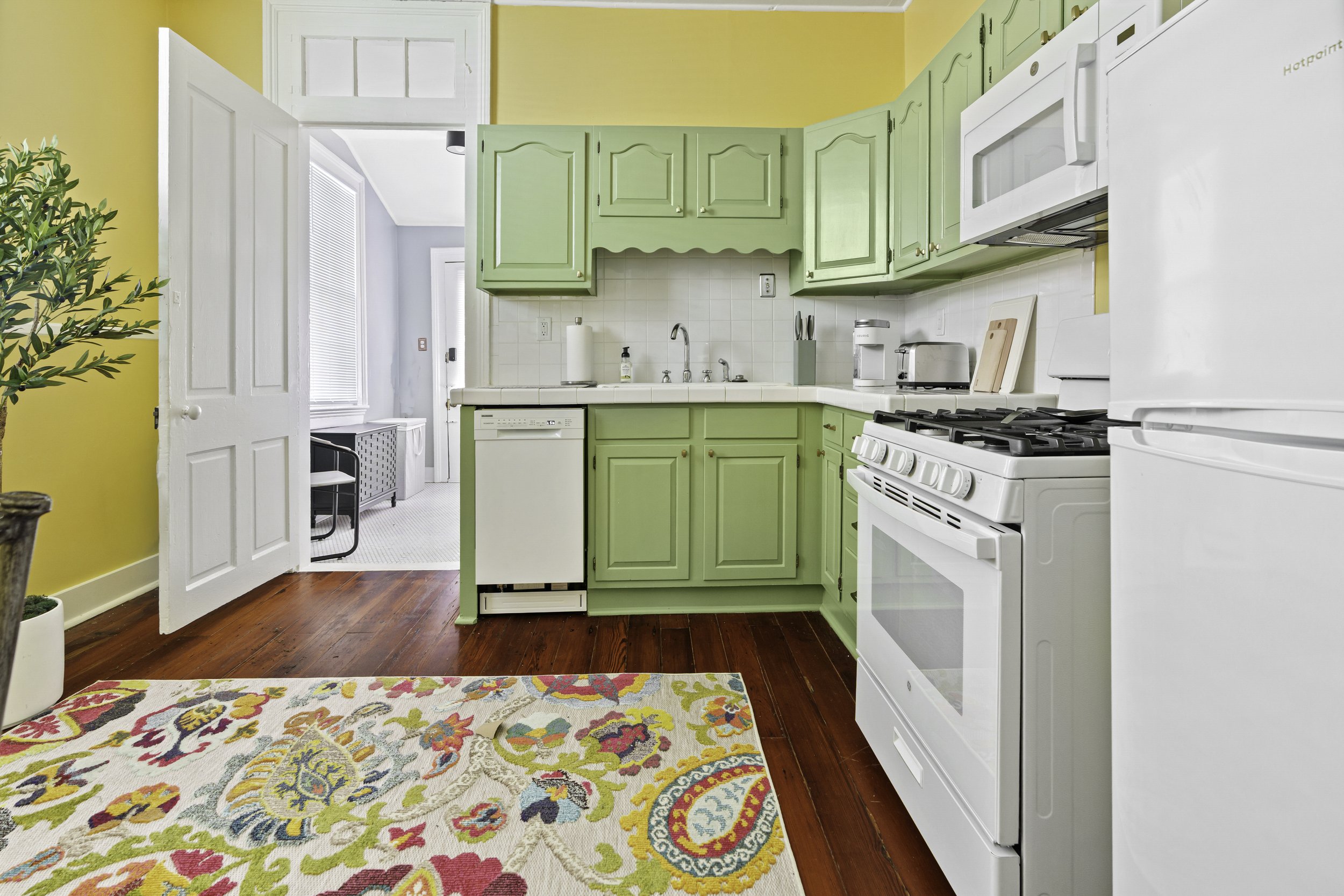 Kitchen with yellow walls and green cabinets, white appliances including a refrigerator, oven, microwave, and dishwasher, hardwood floor, colorful floral rug, and a doorway leading to a room with blue walls and a window.