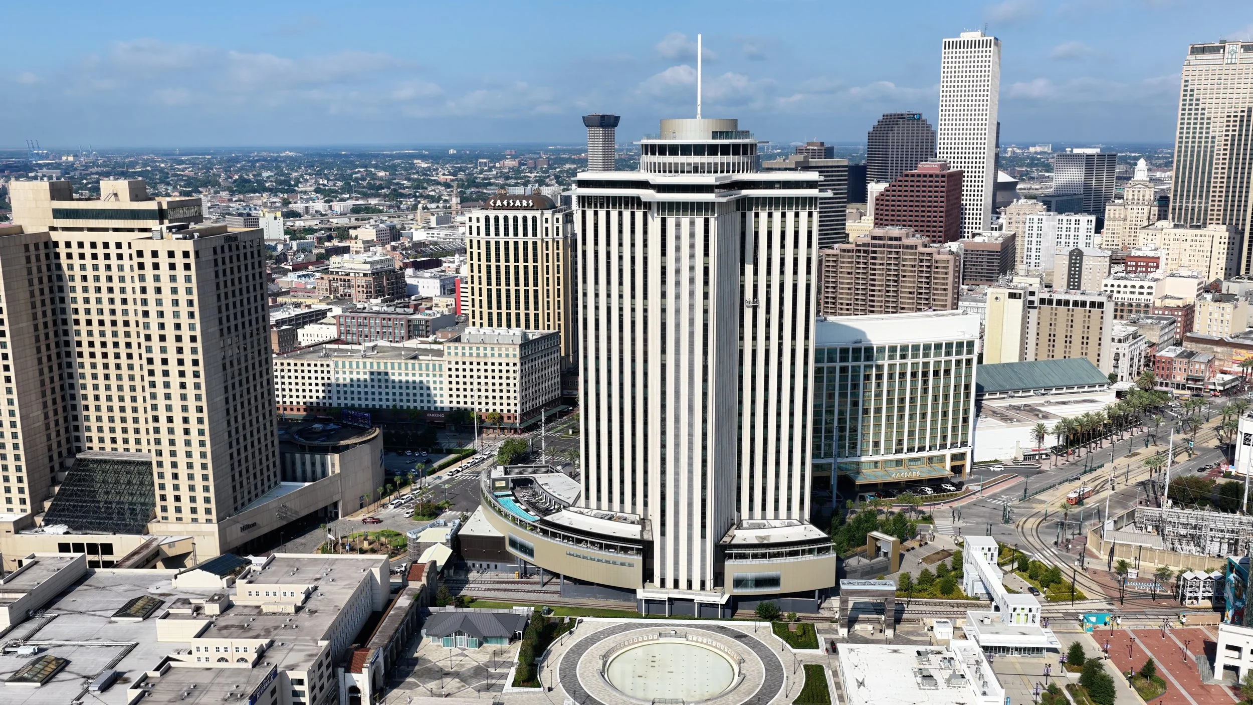 A city skyline with tall skyscrapers, including the Caesars building, under a partly cloudy sky. Urban streets, parking lots, and a roundabout are visible in the foreground.