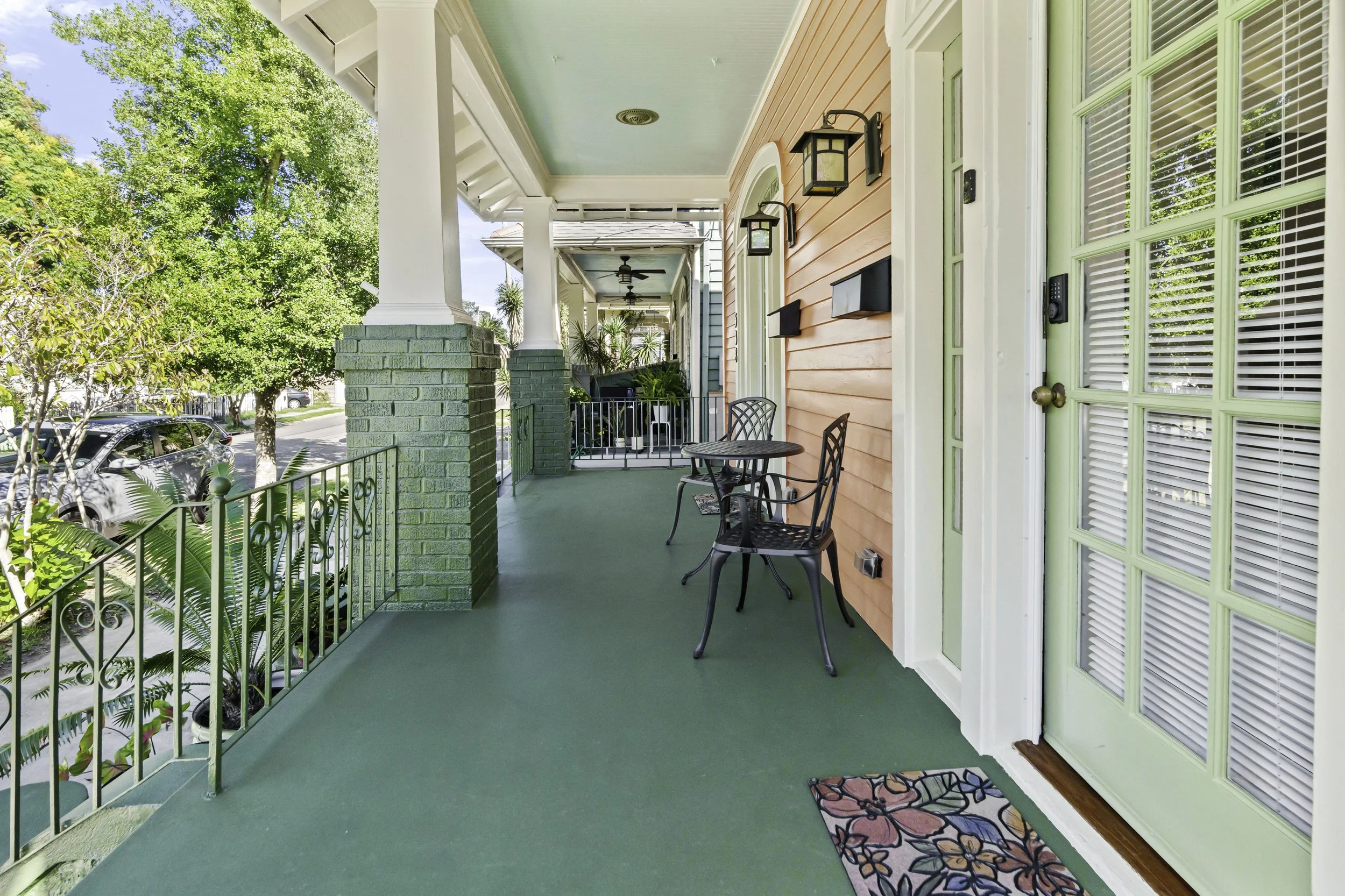 Front porch of a colorful house with a green painted floor, orange walls, and a green front door with white trim, decorated with a floral doormat, black outdoor table, and two black chairs, with a brick and metal railing and lush greenery outside.