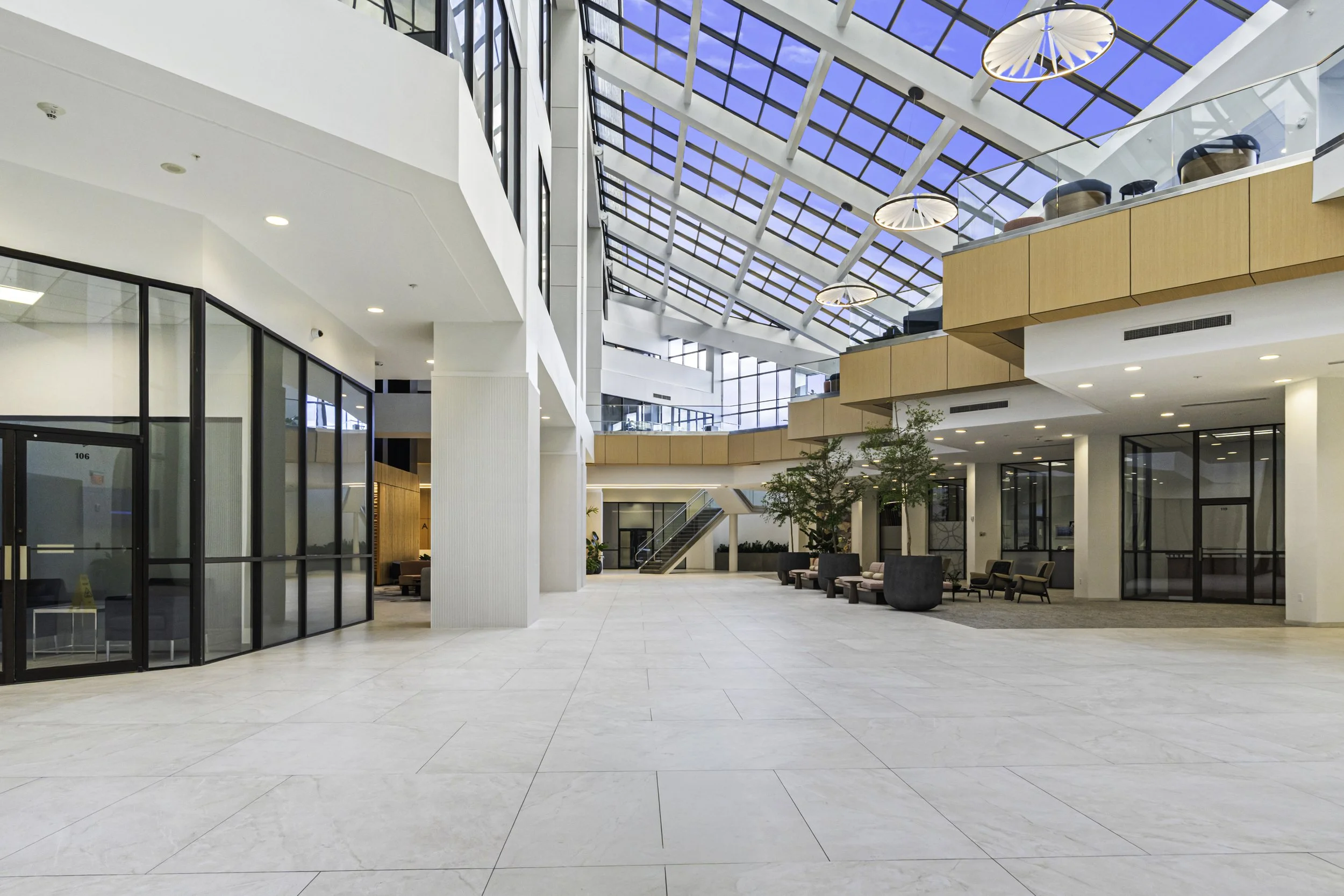 Open lobby area in a modern building with natural light streaming through a large glass ceiling, featuring seating arrangements, a staircase, potted plants, and glass walls.