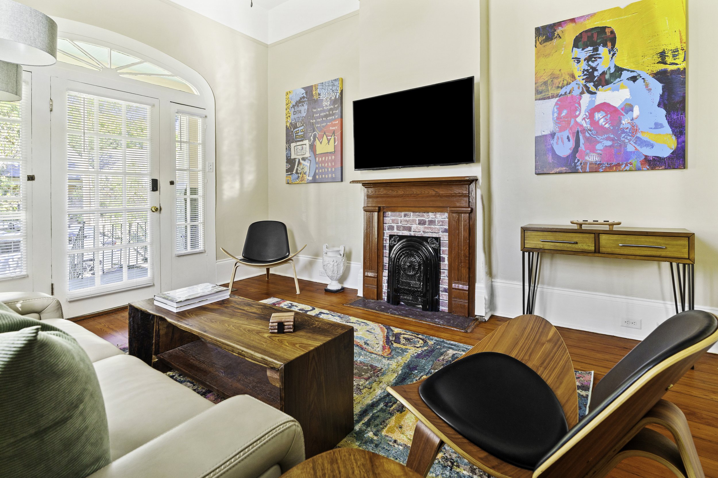 Living room with wooden floor, colorful abstract rug, white sofa with green pillows, two black and wood chairs, wooden coffee table, fireplace with brick and wood mantel, flat-screen TV above fireplace, colorful portraits on the wall, glass doors wit