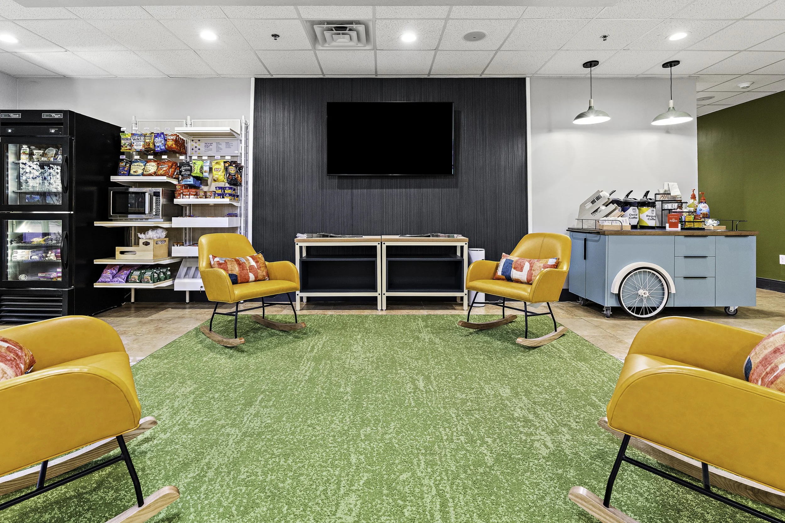 A lounge area with yellow rocking chairs on a green carpet, a black wall with a large blank TV screen, snack shelves, a vending machine, and a coffee station with supplies, pendant lights, and a green wall in the background.