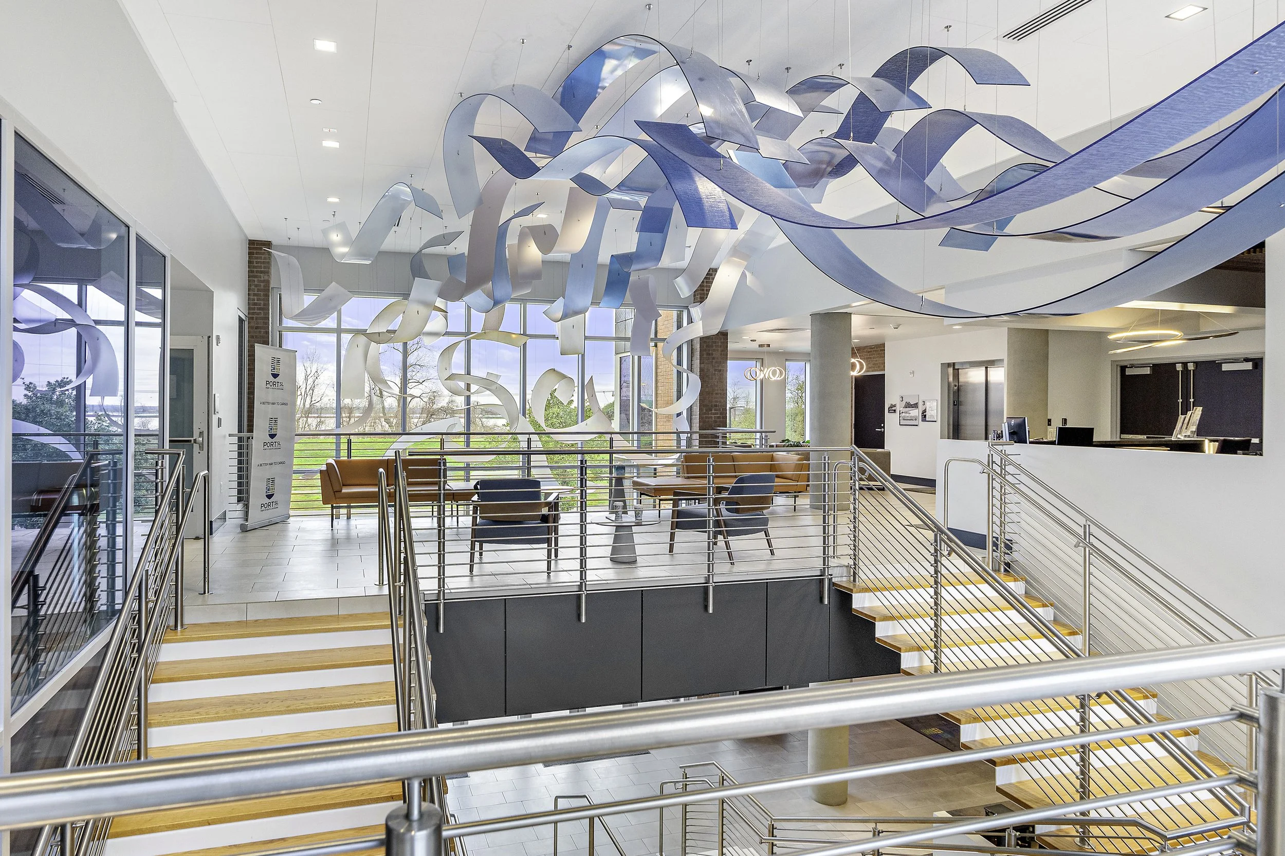 Interior of a modern building featuring a large abstract ceiling installation made of metallic and blue ribbon-like structures, with seating areas, glass walls, and a staircase with metal railings.