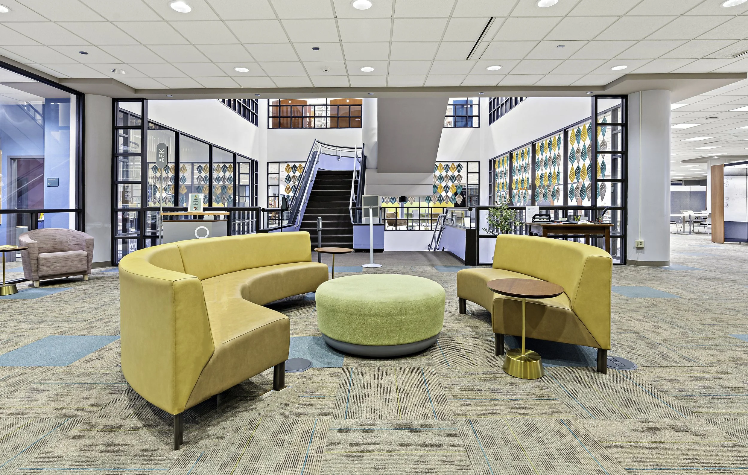Interior view of a modern, open lounge area with colorful seating, including yellow and pink chairs, and a circular ottoman in the center. The space features large glass windows, a staircase, and decorative wall art with leaf patterns.