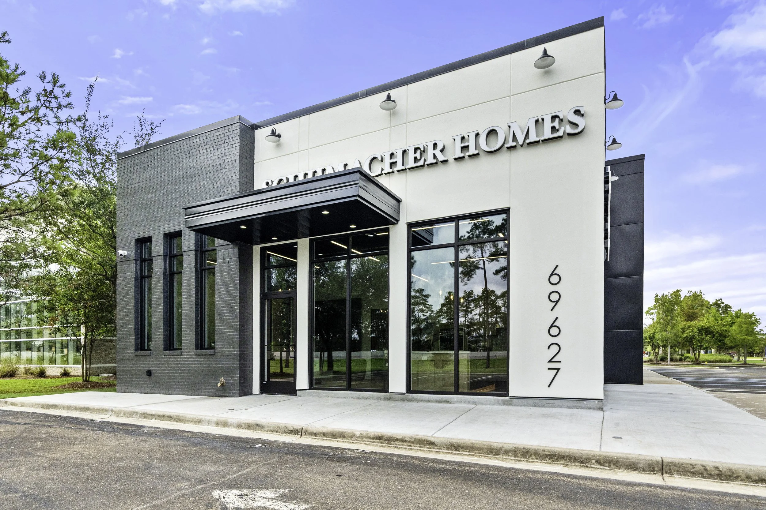 Exterior of a modern townhouse with white and grey facade featuring large glass windows and a black entrance canopy, with the house number 6922 displayed vertically on the side, surrounded by trees and a driveway.