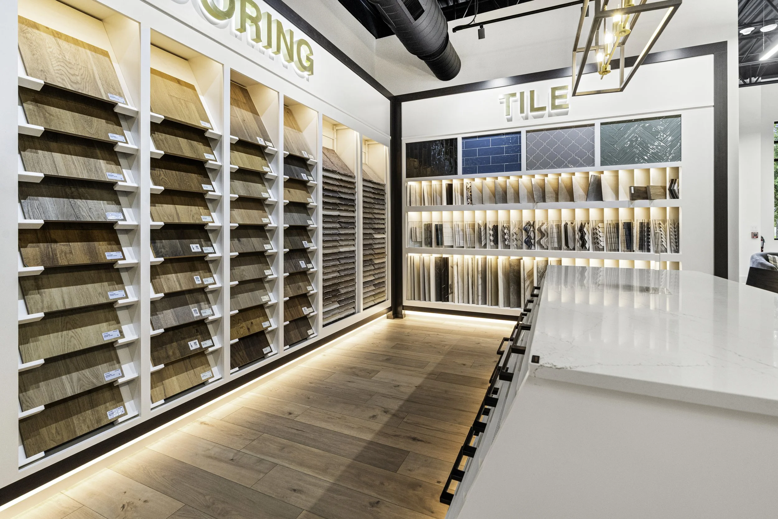 A showroom displaying various wood flooring samples on white shelves labeled 'FLOORING' and 'TILE', with light wood flooring on the floor and a modern lighting fixture on the ceiling.