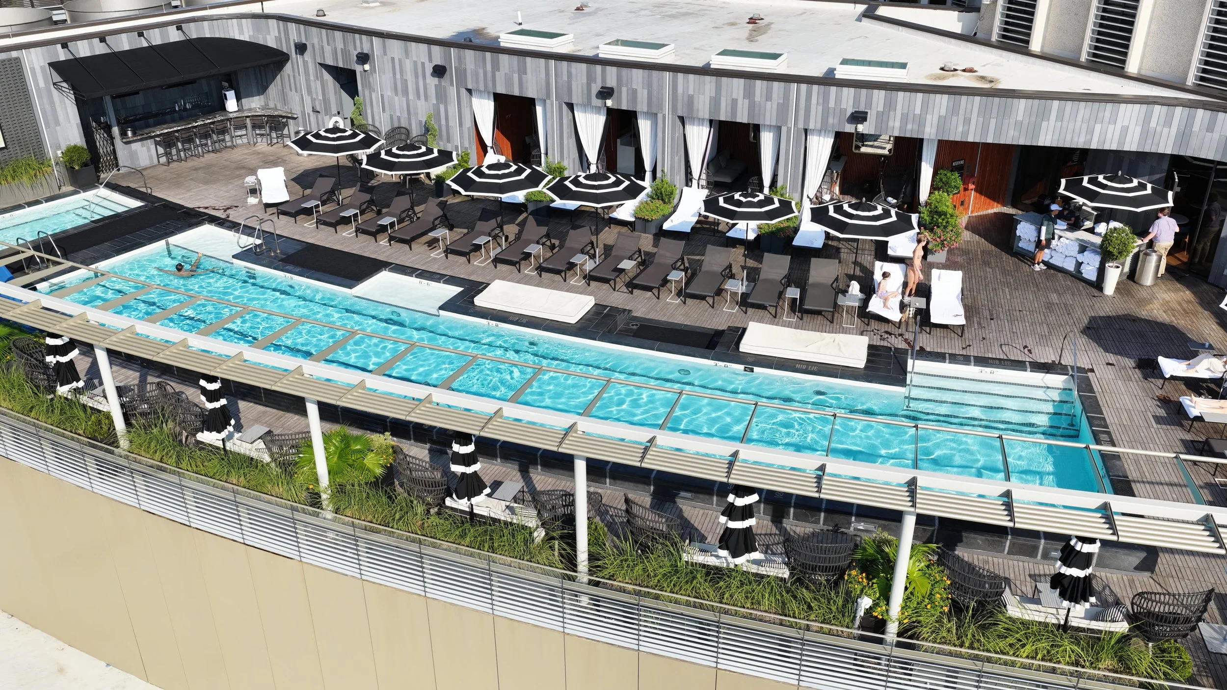 A rooftop swimming pool area with surrounding lounge chairs and umbrellas, some people relaxing and swimming, with a building exterior in the background.
