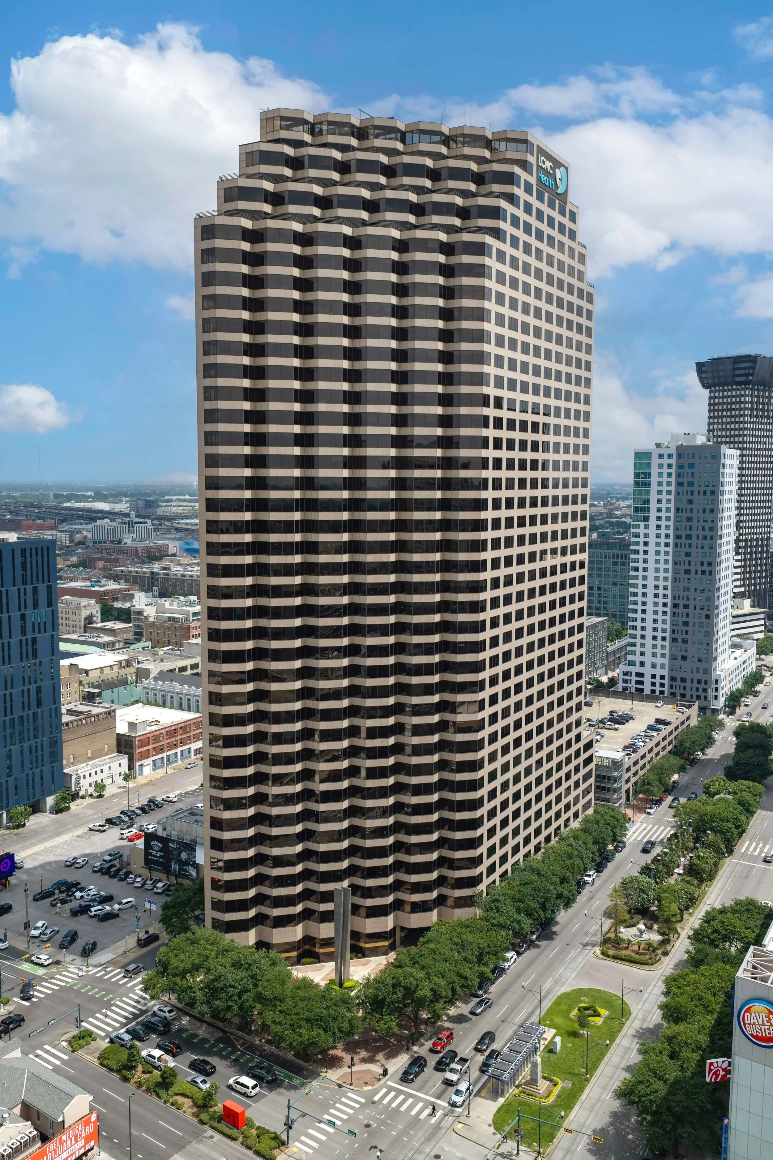A tall office building with a distinctive zigzag pattern on its exterior taken from an aerial view. The skyline features other high-rise buildings, and streets below are busy with cars and pedestrians, framed by green trees and city infrastructure un