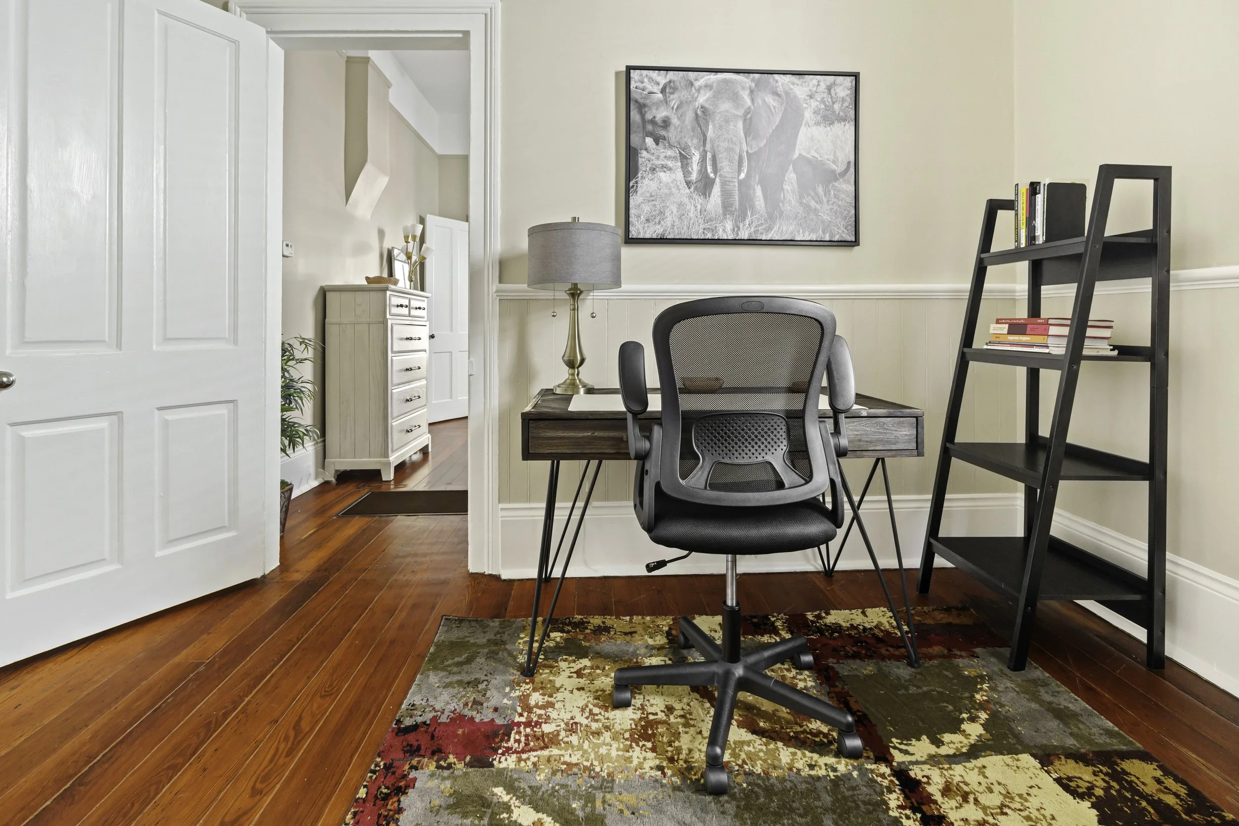 Home office with desk, ergonomic chair, black bookshelf, framed elephant photo on wall, standing lamp, hardwood floors, and decorative rug.