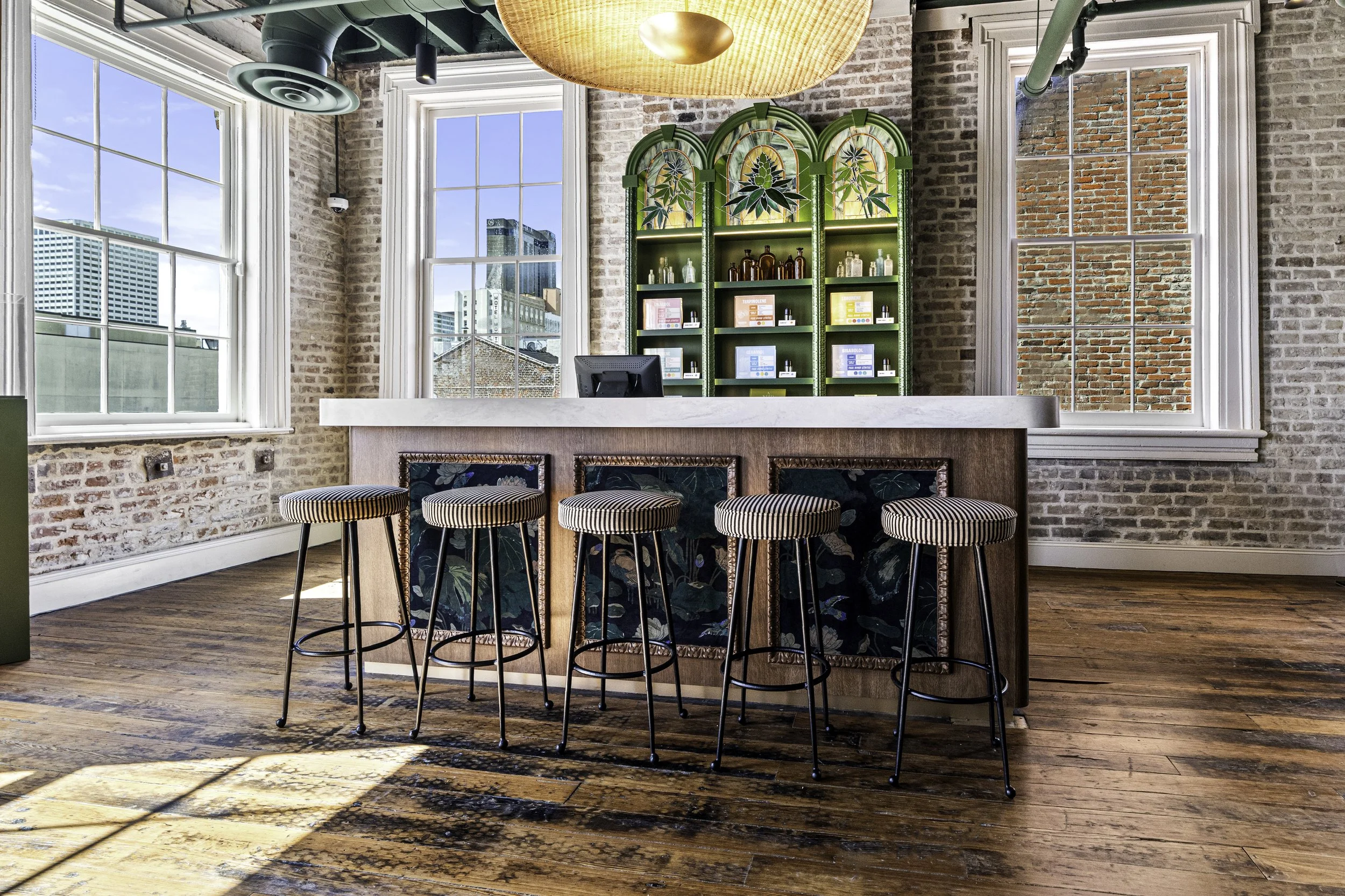 Interior of a brick-walled cafe or bar with large windows, a white marble bar counter, and bar stools with striped padding and floral fabric backs. A decorative green shelving unit with stained glass and botanical designs is behind the counter. There
