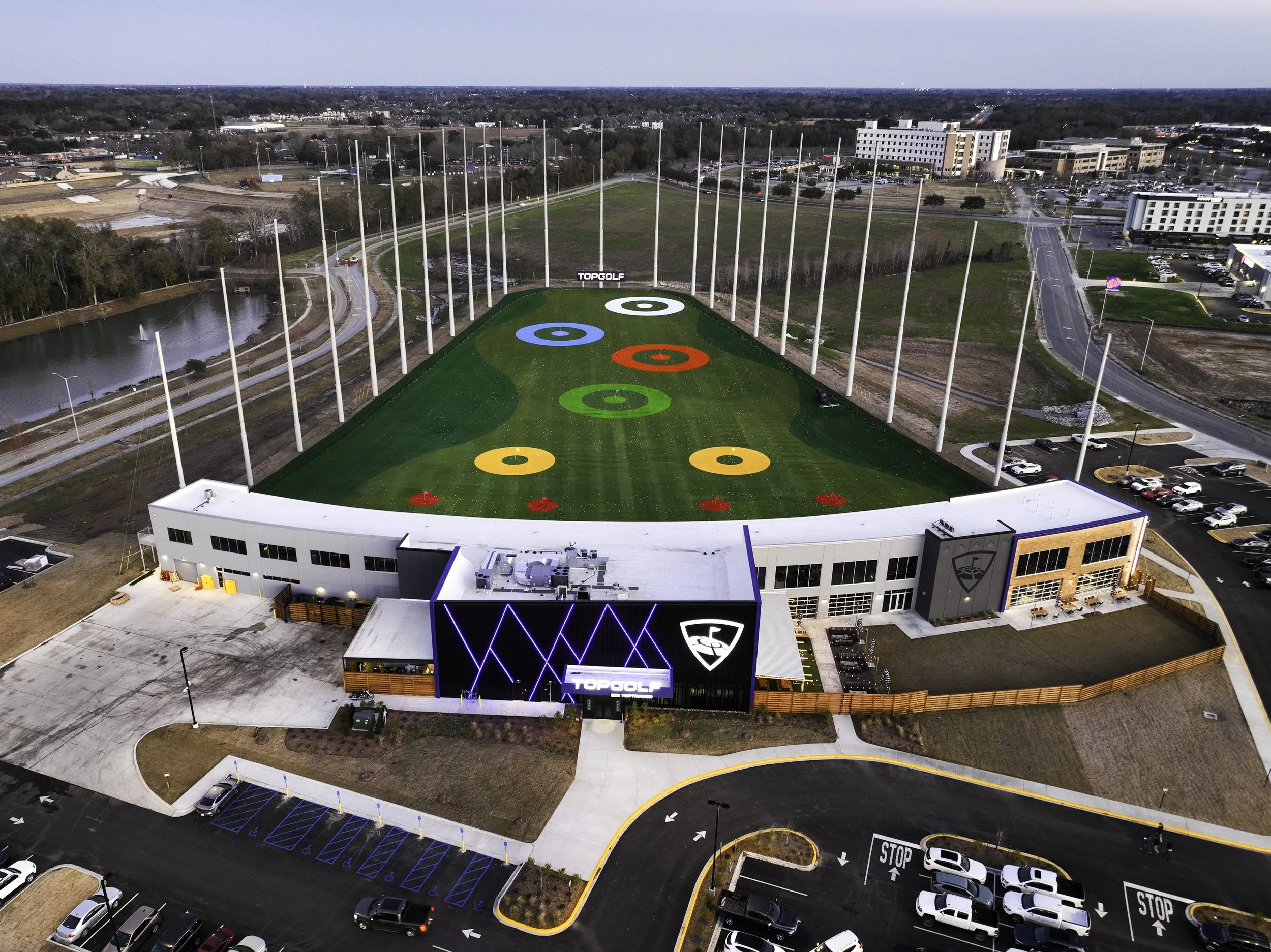 Aerial view of a golf driving range with colorful targets, surrounding parking lots, roads, and nearby buildings.