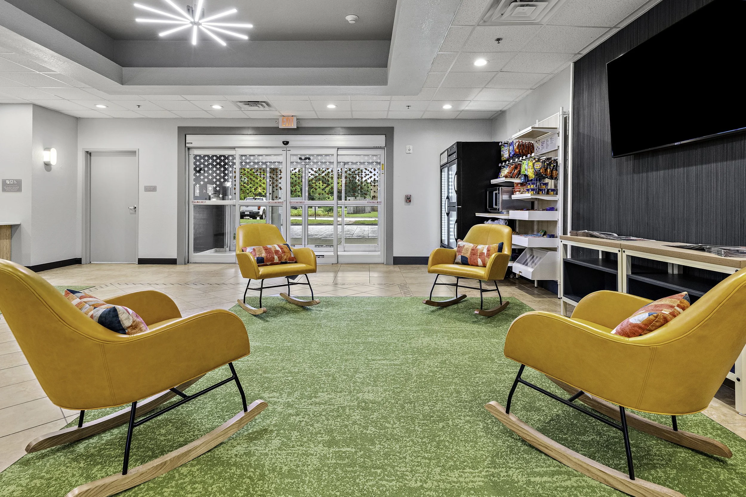 A spacious indoor lobby with orange rocking chairs, a green rug, a vending machine, a snack shelf, a large TV screen, and glass doors leading outside.