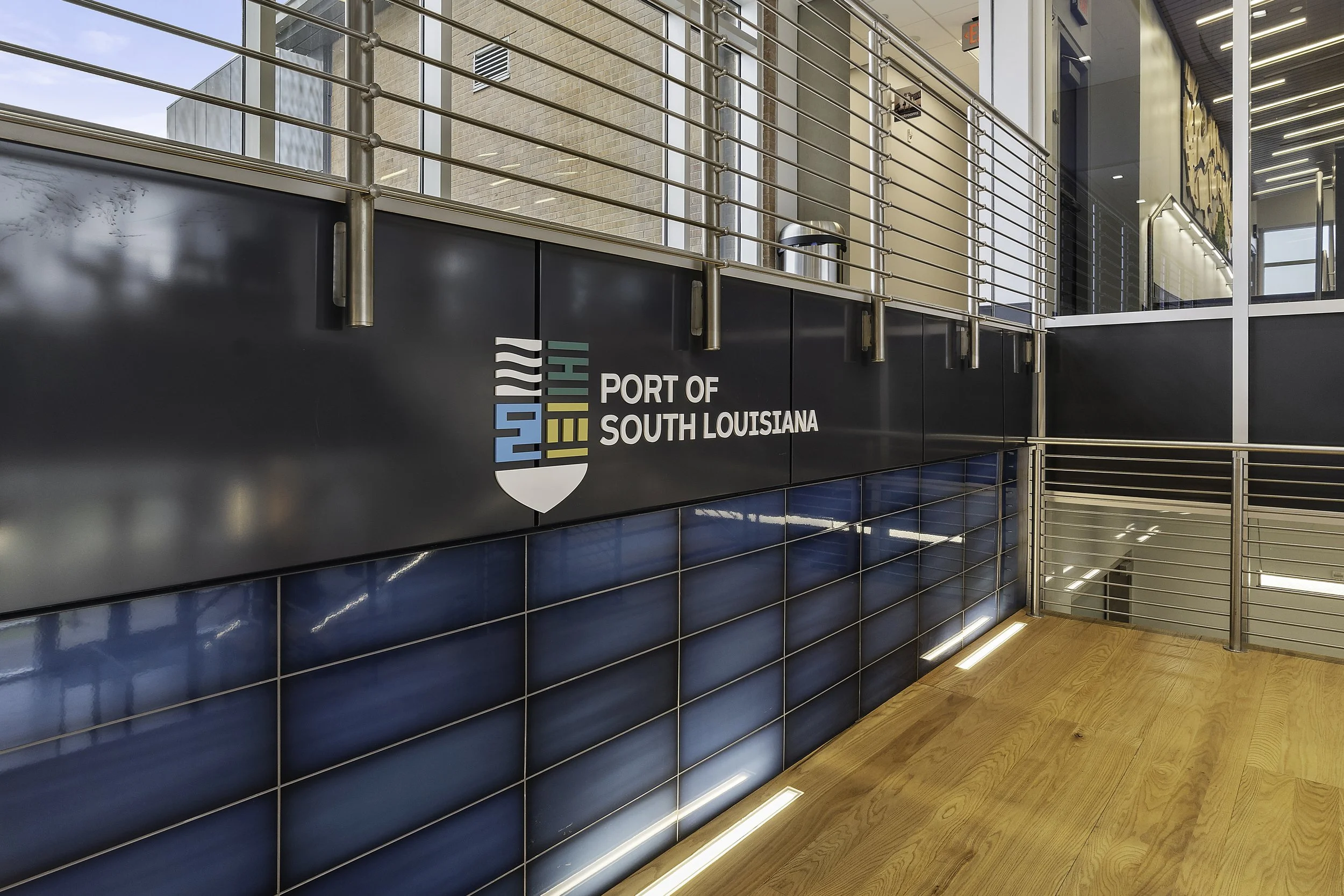 Interior of a modern building with a sign reading 'Port of South Louisiana,' metal railing, blue tiled wall, and wooden flooring.