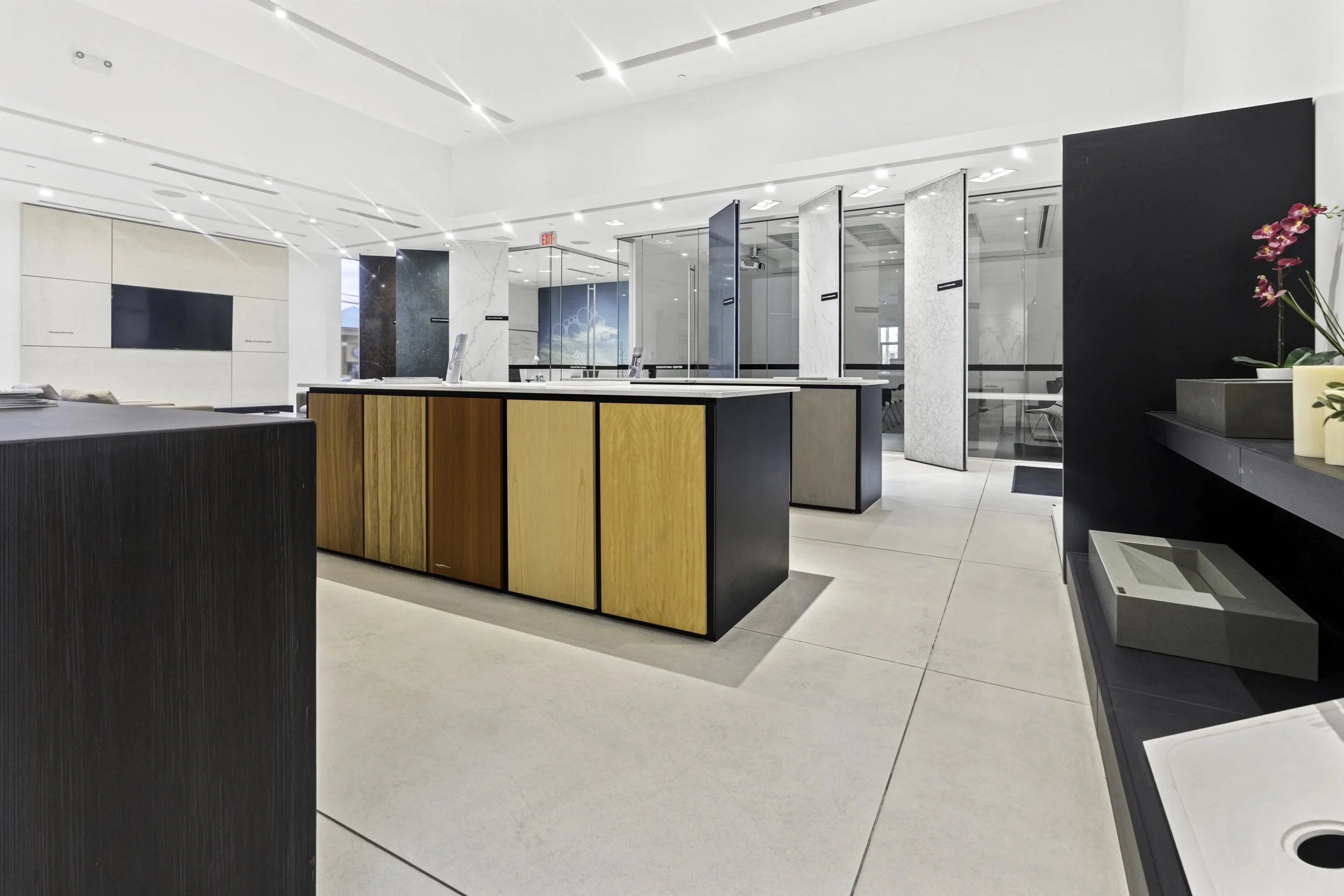 Modern interior space with a service counter featuring wood panels, glass partitions, white walls, and ceiling lights, with an orchid plant on a dark gray counter to the right.