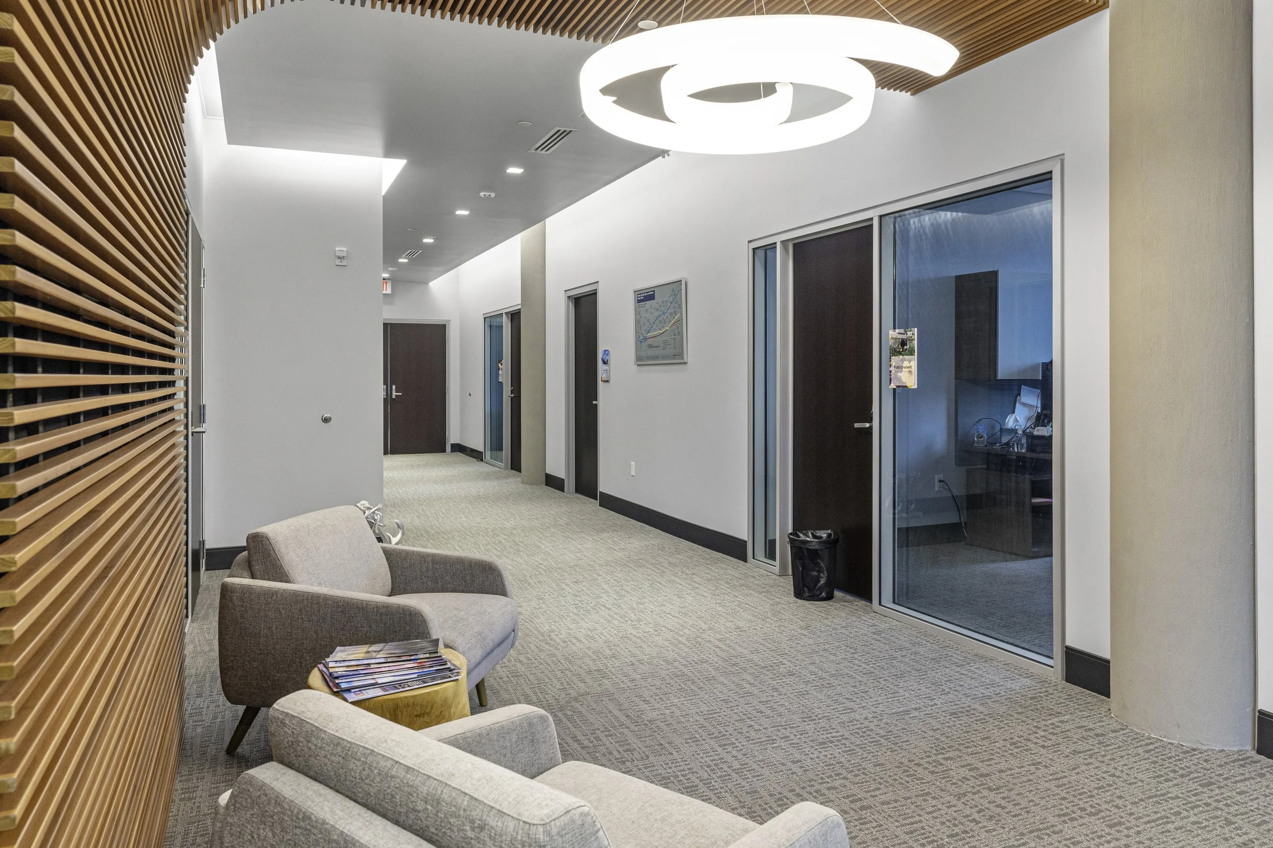 An office corridor with beige carpet, white walls, and a wooden slatted wall on the left. There are modern ceiling lights and a spiral-shaped light fixture. On the right, glass-enclosed rooms with dark doors, one with a visible computer and cables in