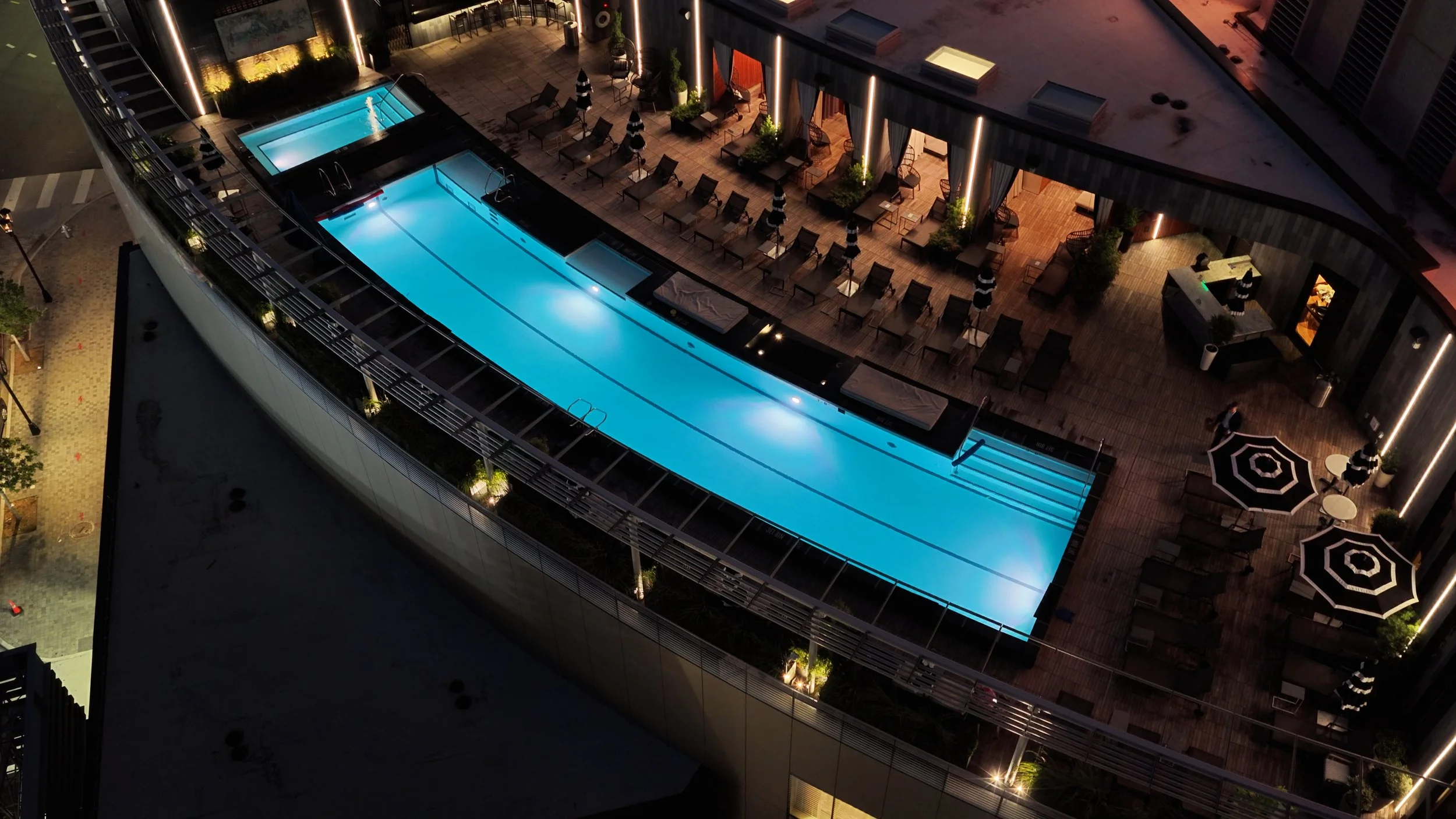 Aerial view of a rooftop pool illuminated at night, surrounded by lounge chairs, umbrellas, and outdoor seating areas with modern lighting.