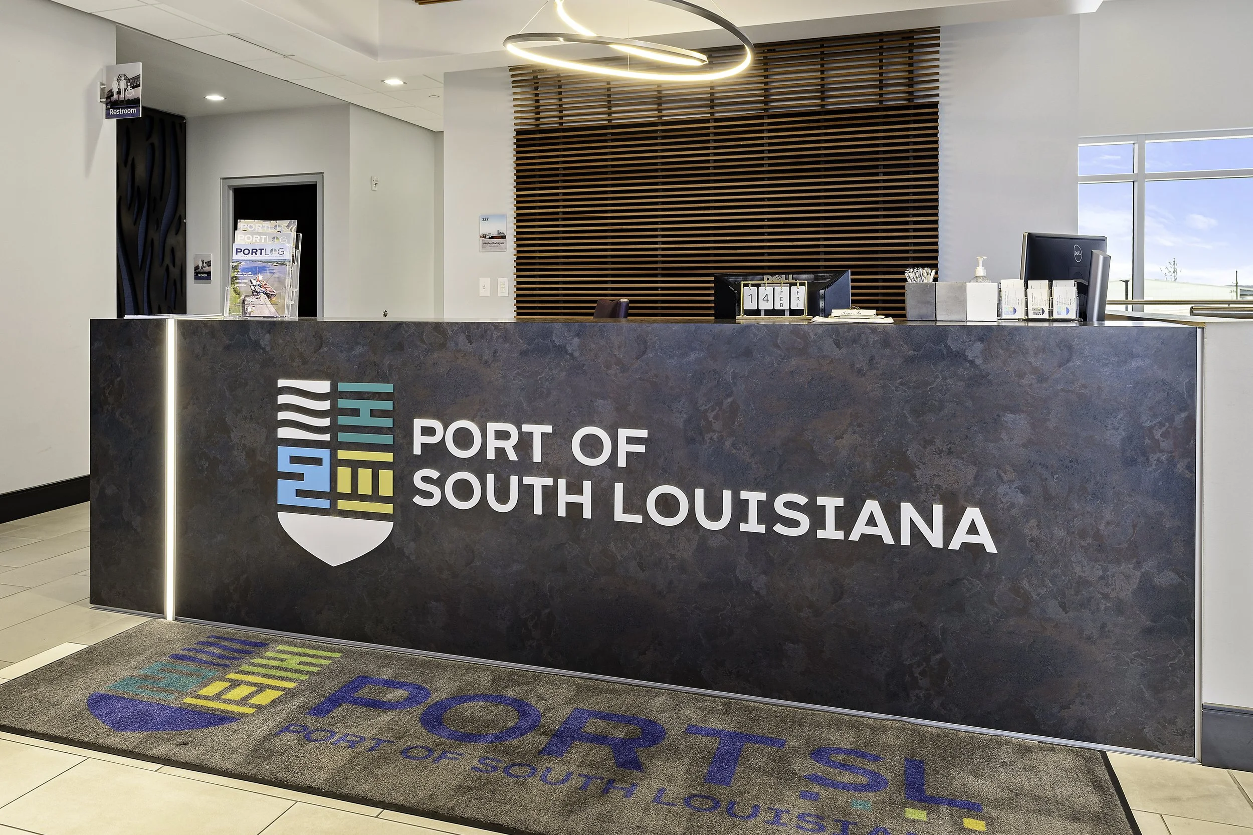 Reception desk at the Port of South Louisiana with a colorful logo, computer, and office supplies. Behind the desk is a wooden slat wall and a large window showing a blue sky. Floor mat with the port's logo is in front of the desk.