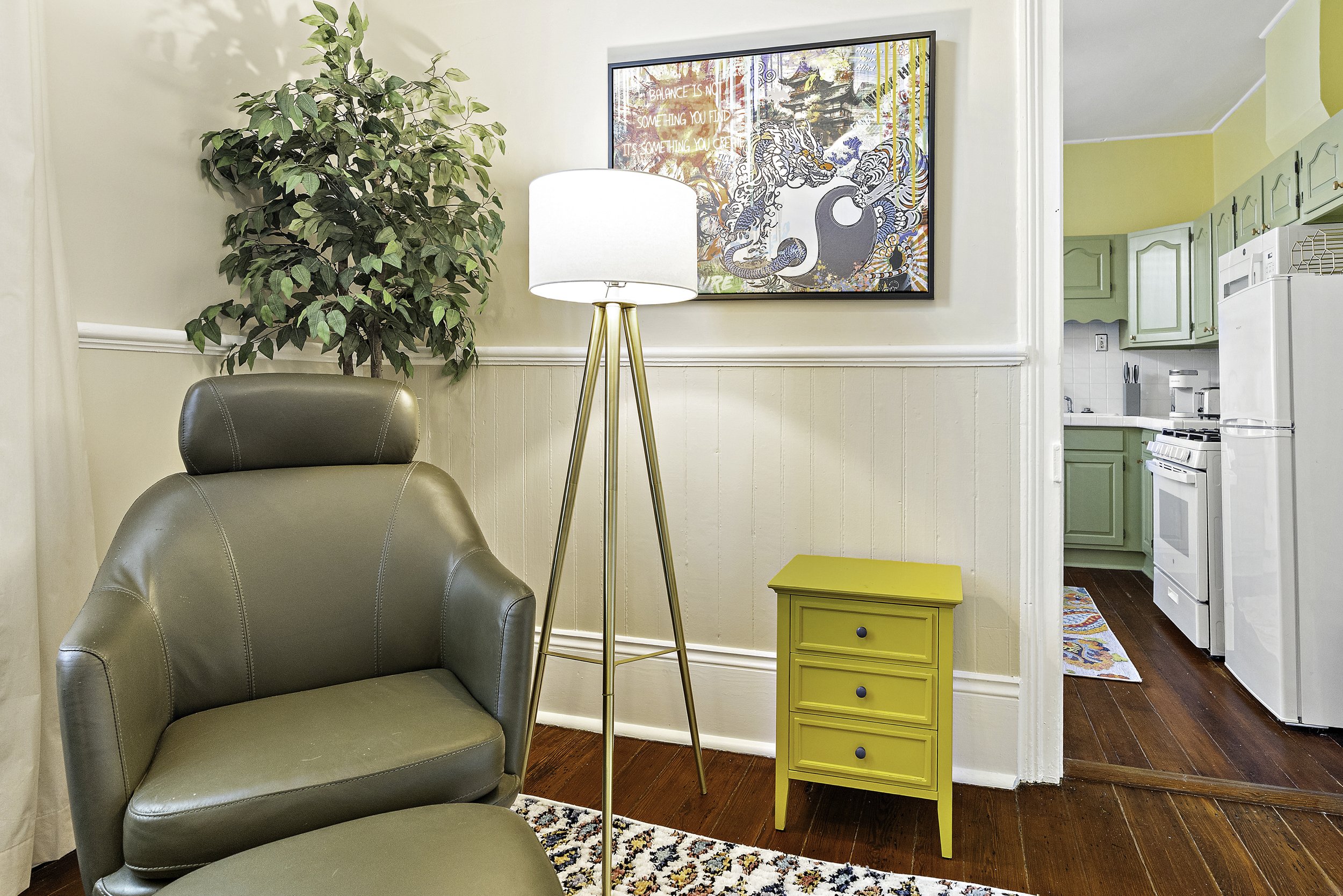 Living room with a gray leather armchair, a yellow side table, a tall floor lamp with a white shade, a large potted plant, a colorful wall art, and a doorway leading to a kitchen with green cabinets and white appliances.
