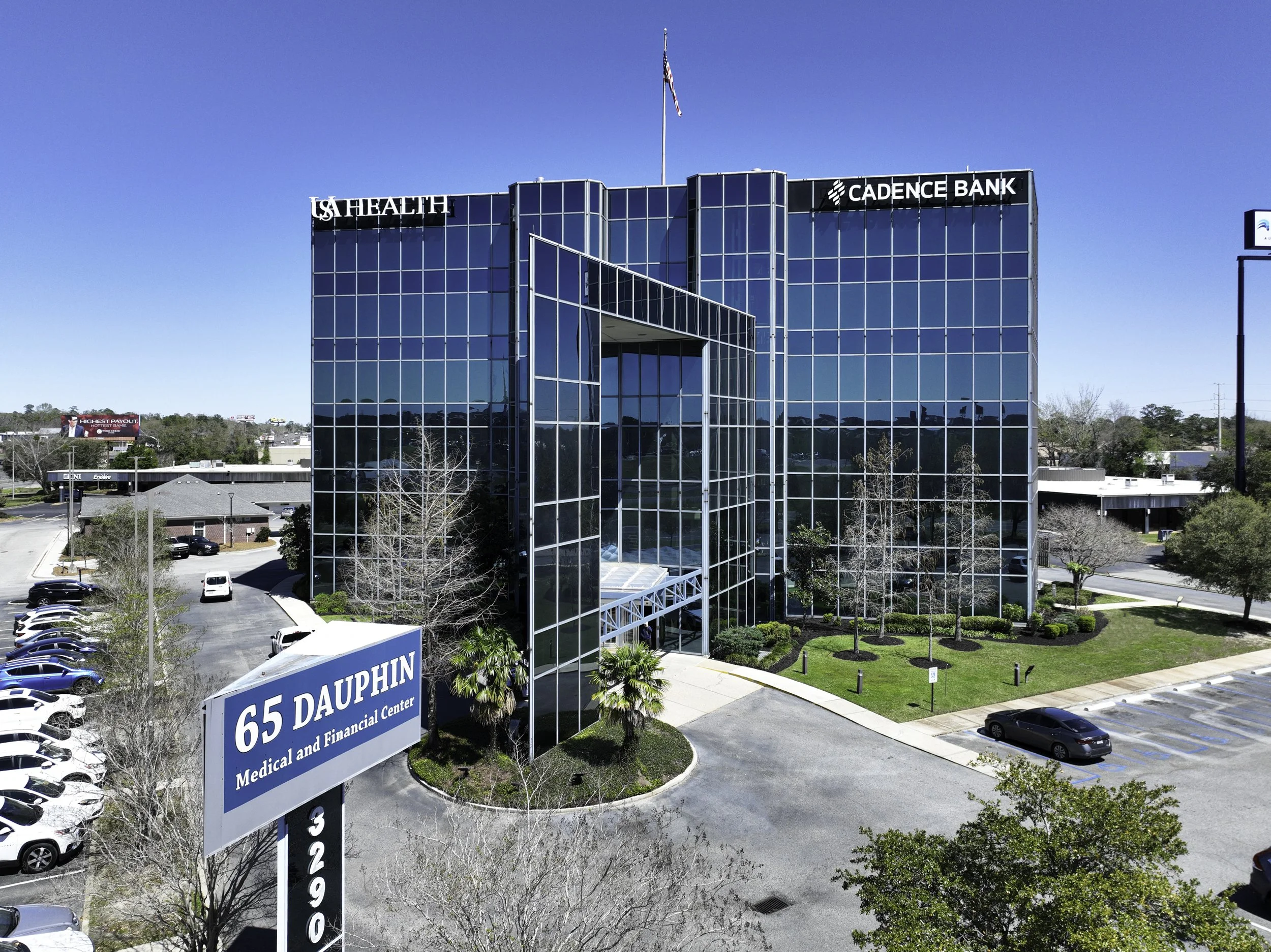 Modern glass office building with signs for LA Health, Cadence Bank, and Dolphin Medical and Financial Center at 65 Dauphin Street. Parking lot with cars, trees, and a clear blue sky.