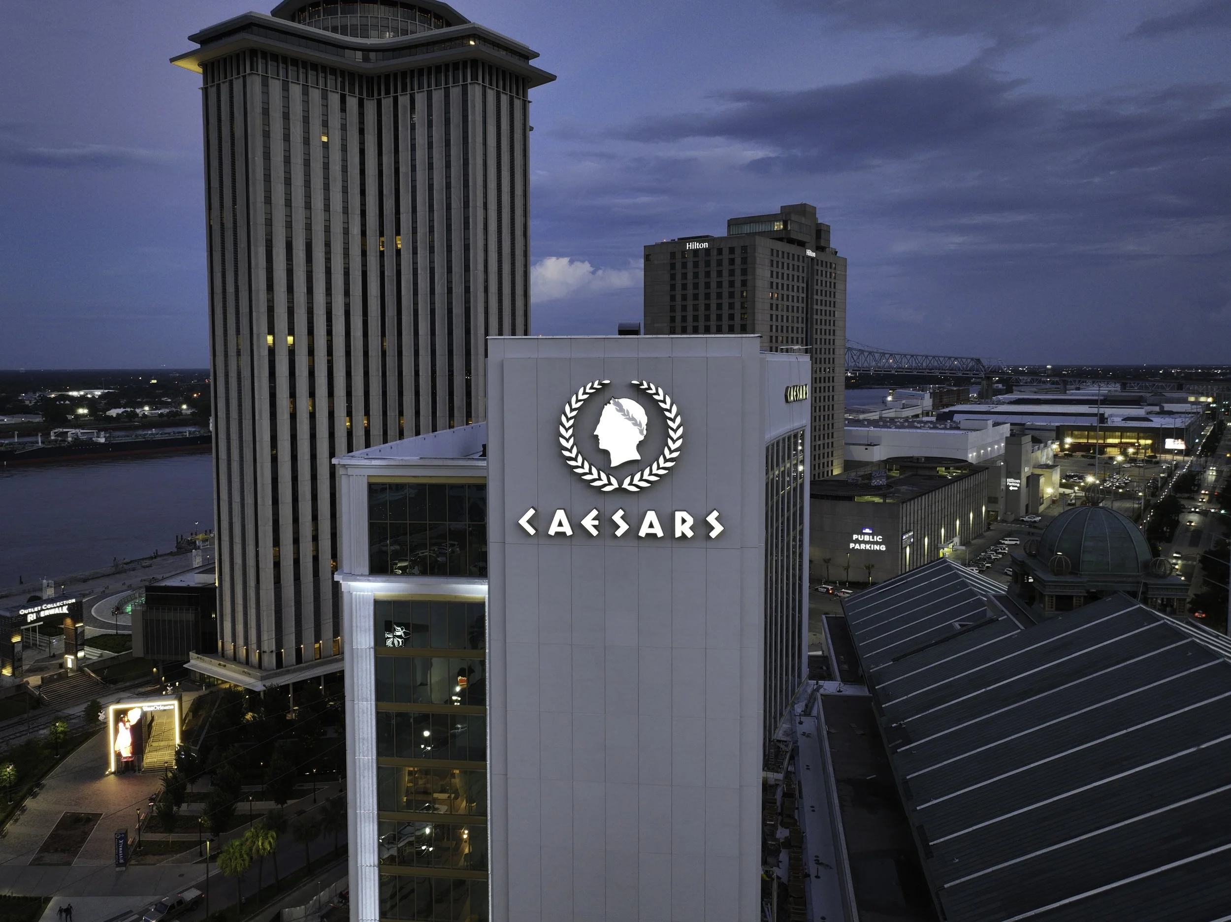 View of a city skyline at dusk featuring tall buildings, including a Caesars building with its logo prominently displayed. The sky is partly cloudy with some lights on in the buildings and street below.