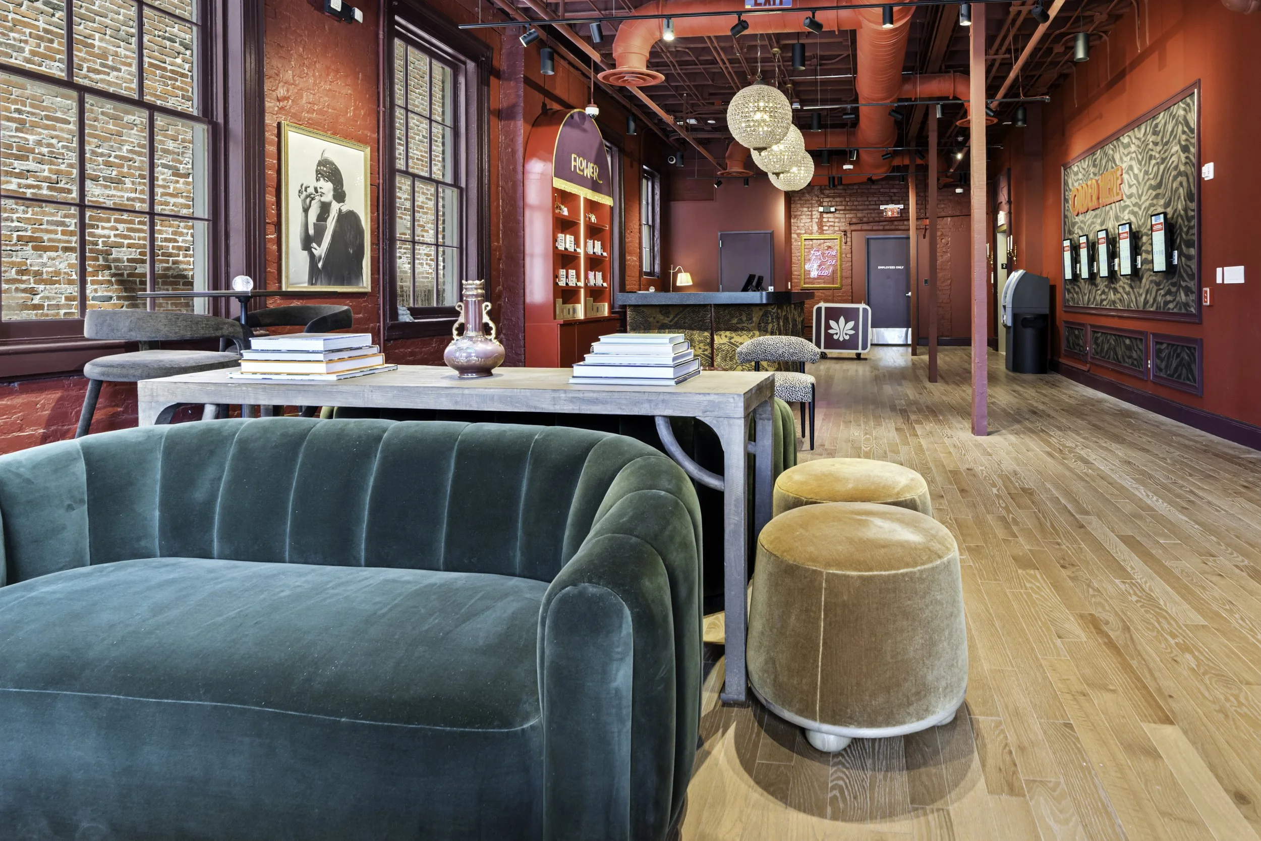 Interior of a modern, stylish lounge or cafe with red walls, large windows, a seating area with green velvet chairs, and tables with magazines. Decor includes artwork, a red bookshelf, and a reception desk in the background.