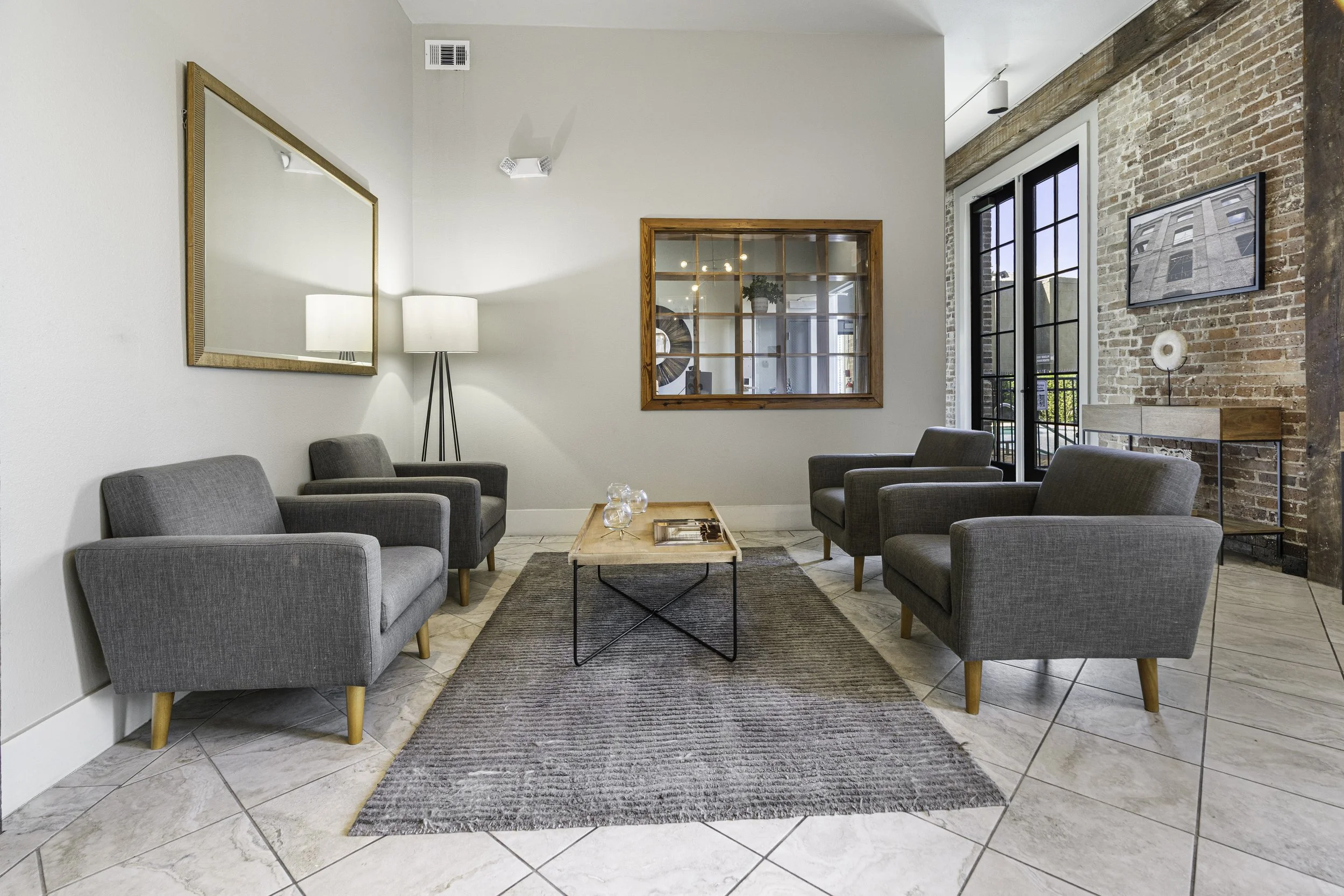 A modern waiting area with four gray armchairs around a small wooden coffee table, a dark rug, a framed mirror, a tall floor lamp with a white shade, large windows with black frames, a brick wall with a framed photo, and a wooden window frame on a wh