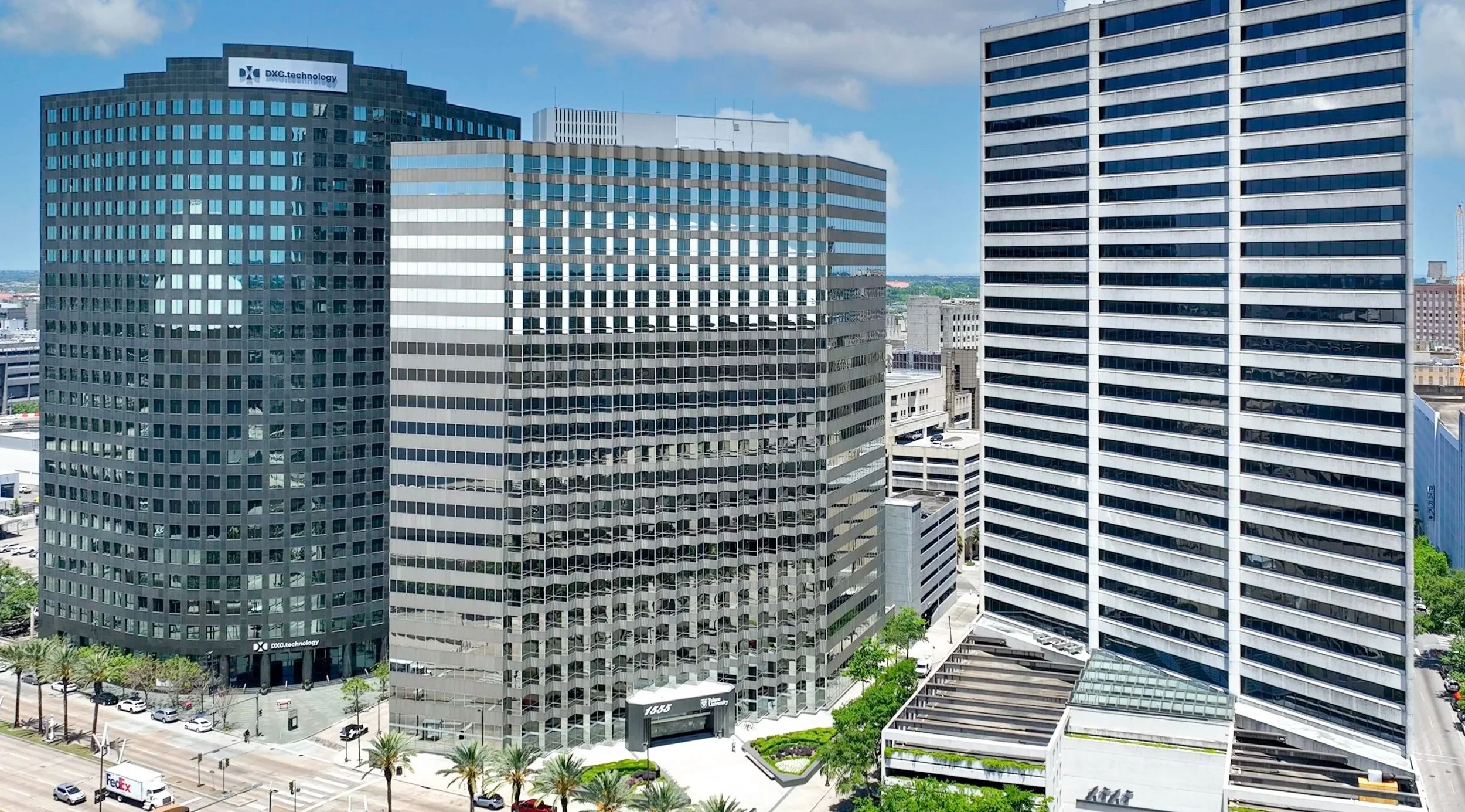 Several tall modern office buildings with glass windows in a cityscape under a partly cloudy sky. Streets and parked cars are visible at the street level.