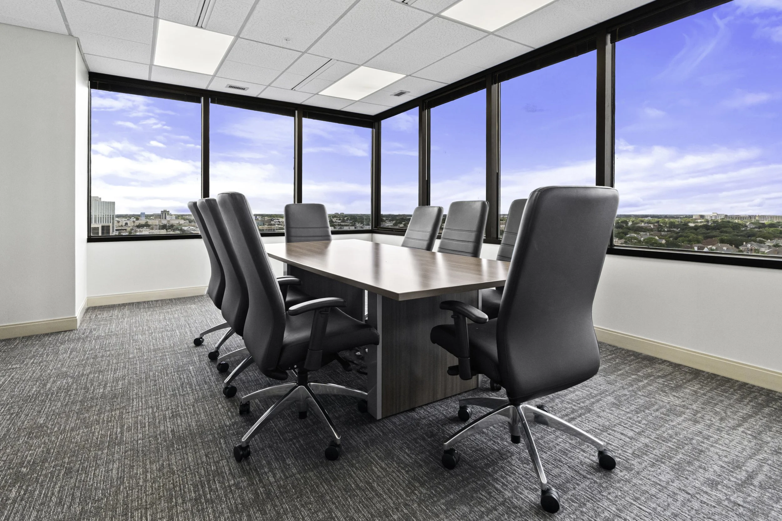 Conference room with a large wooden table surrounded by black leather office chairs, large windows revealing a city skyline under a blue sky with clouds.