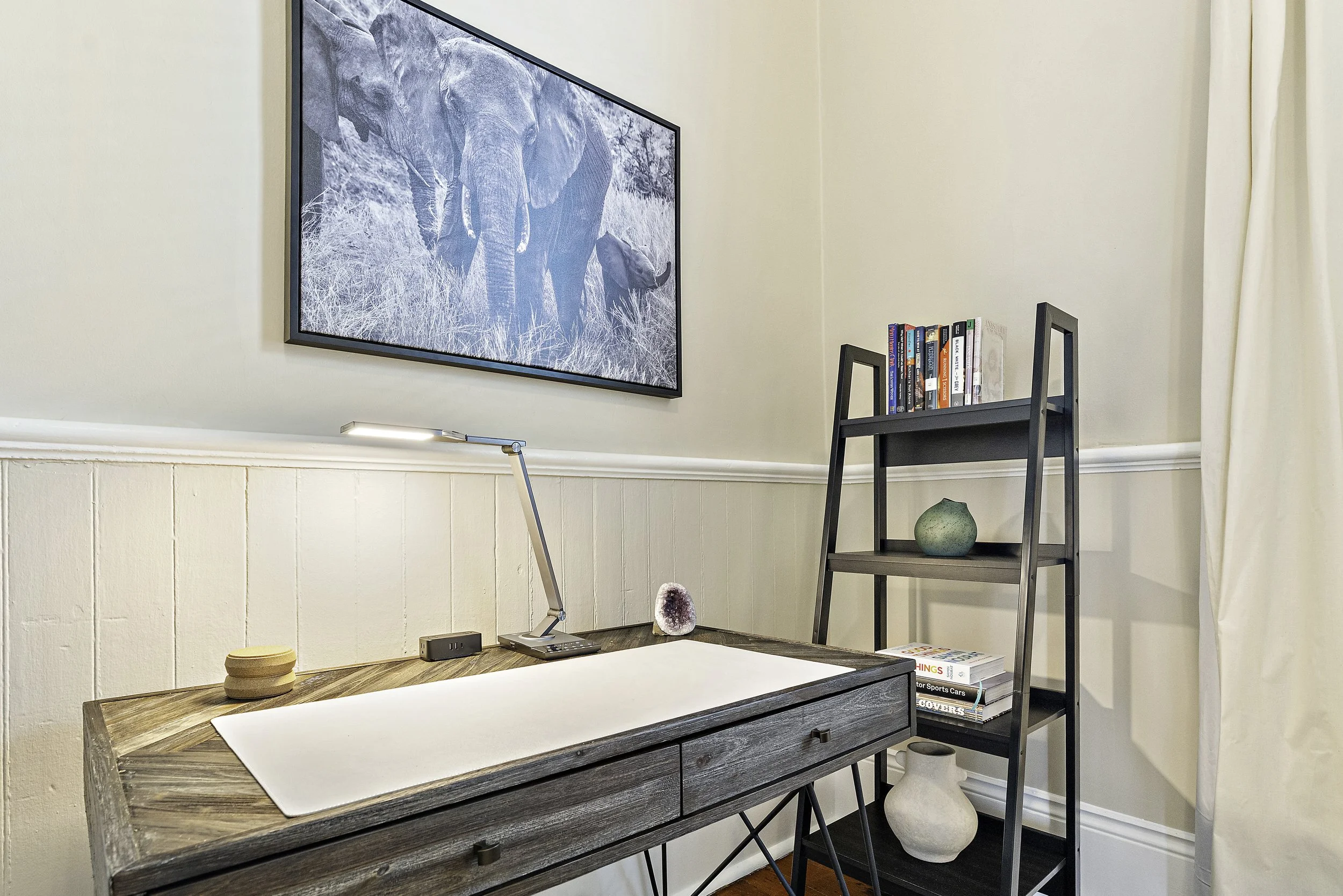 Home office with a wooden desk, a white mat or pad, a modern desk lamp, and a small decorative stone. A wall-mounted TV displays an elephant image, and a black ladder-style shelf holds books and decorative vases in a corner of the room.