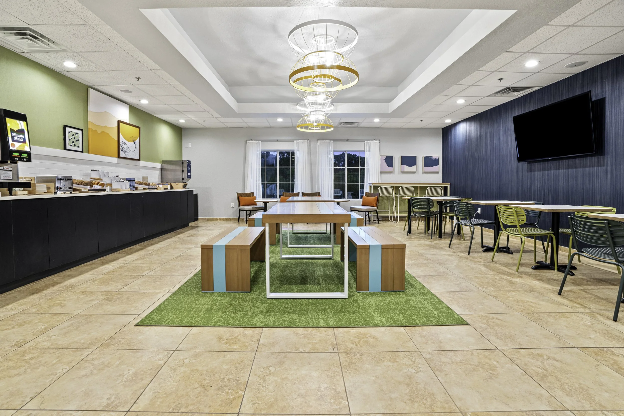 A bright indoor cafe or lounge with modern decor, featuring a green area rug, tables, chairs, and a ceiling with stylish light fixtures, window curtains, artwork on the walls, and a television mounted on a blue textured wall.