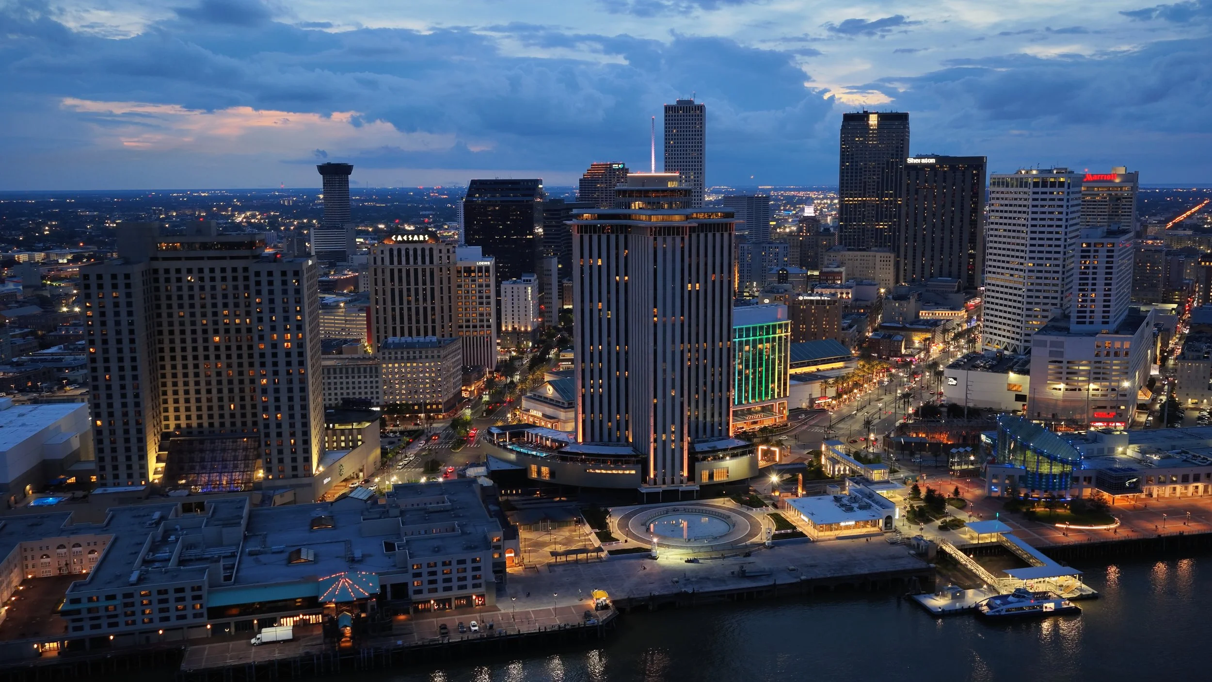 A city skyline at dusk with tall buildings, some illuminated, and a waterway in the foreground.