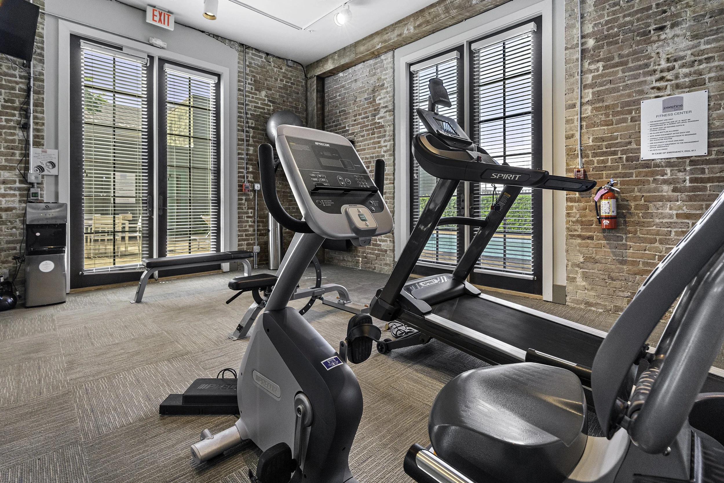 Interior view of a gym with brick walls, large windows with blinds, and exercise equipment including a treadmill and an elliptical machine, with an emergency exit sign on the ceiling.