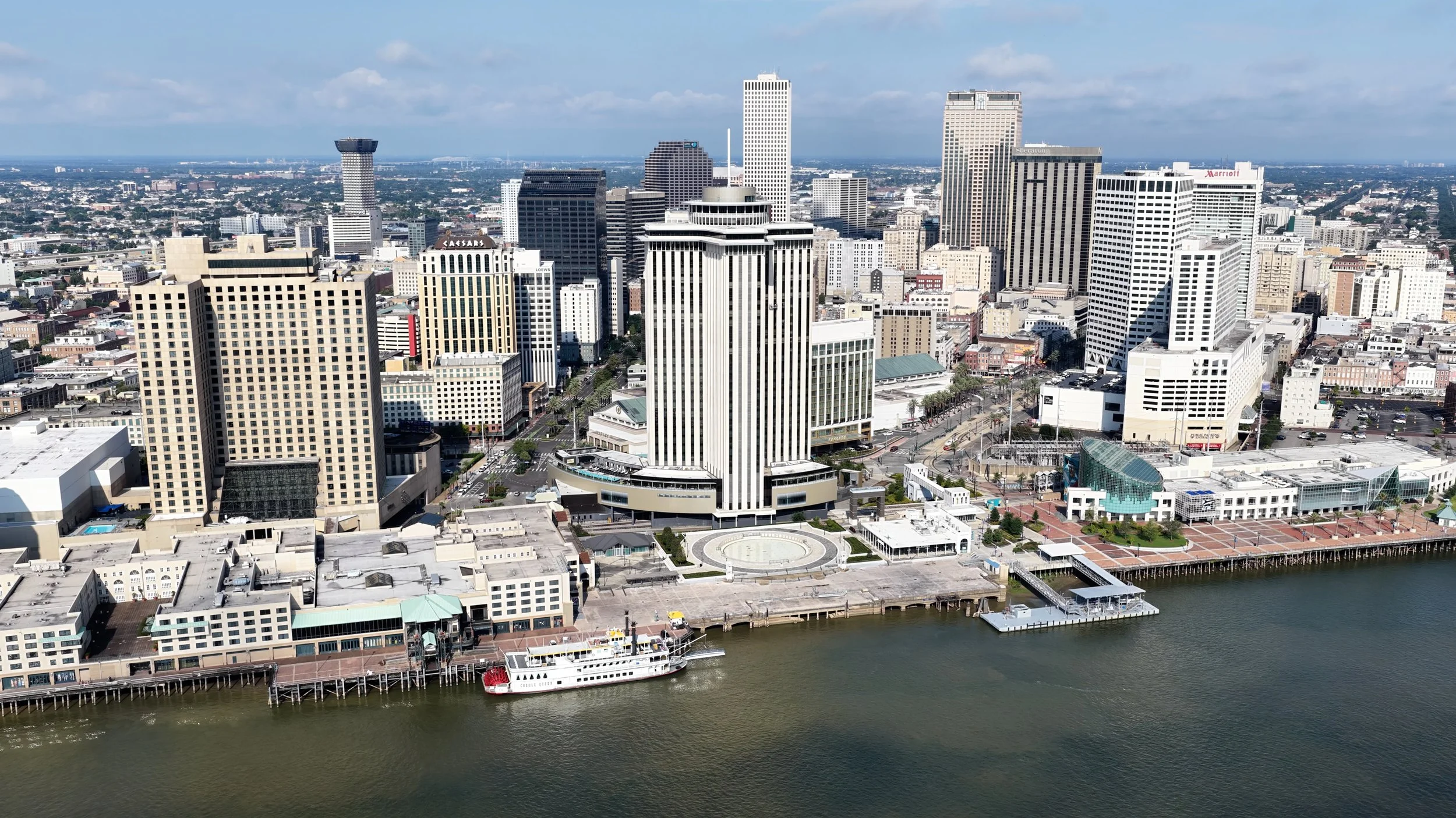 Aerial view of a city skyline with tall buildings, a waterfront with boats, and a river in the foreground.