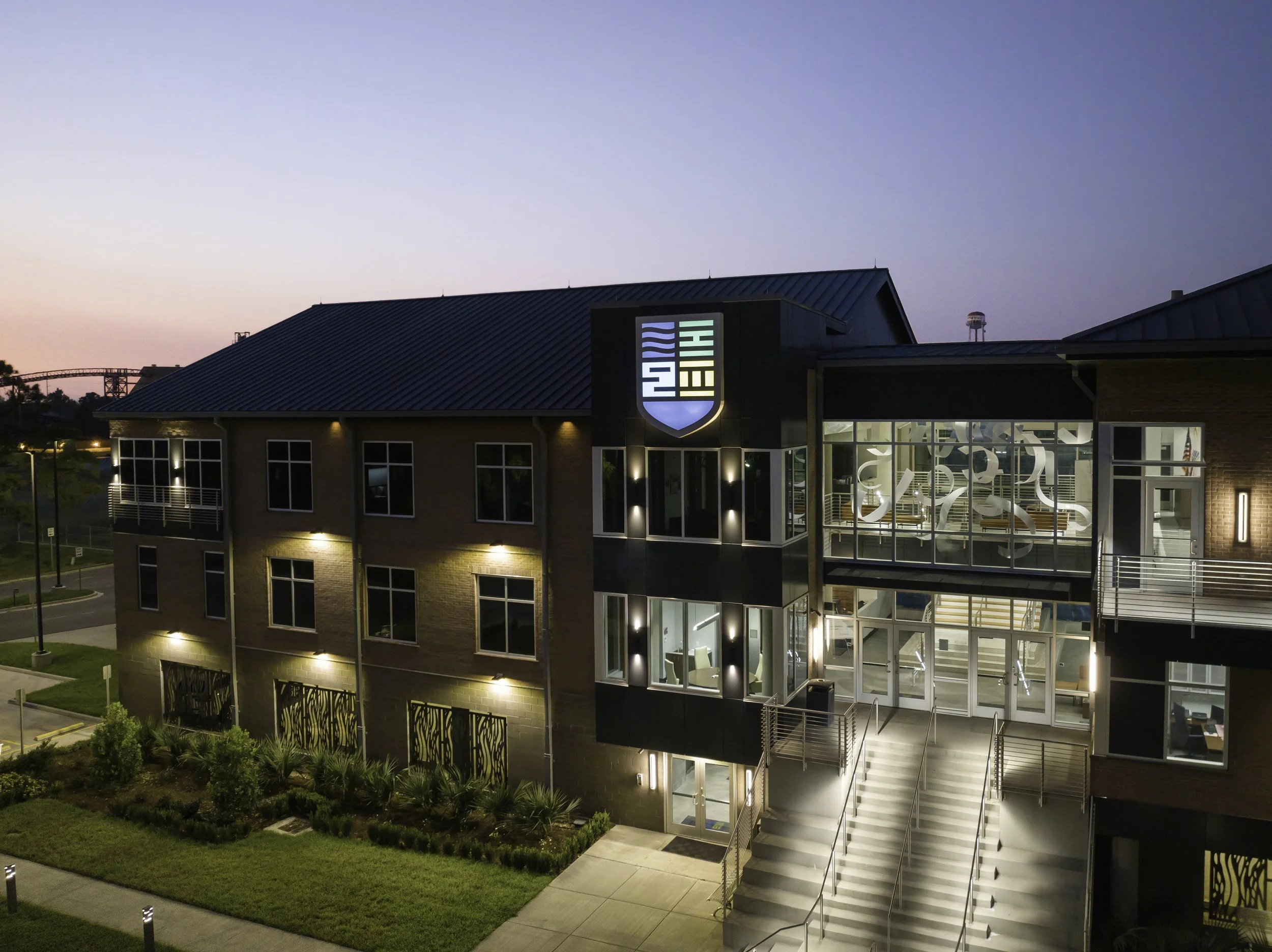 A modern multi-story building during twilight with illuminated interior and exterior lights, featuring a sign that says "HIRE" on the front and a glass entrance with stairs leading up to it, surrounded by landscaped greenery and small lighting fixtur