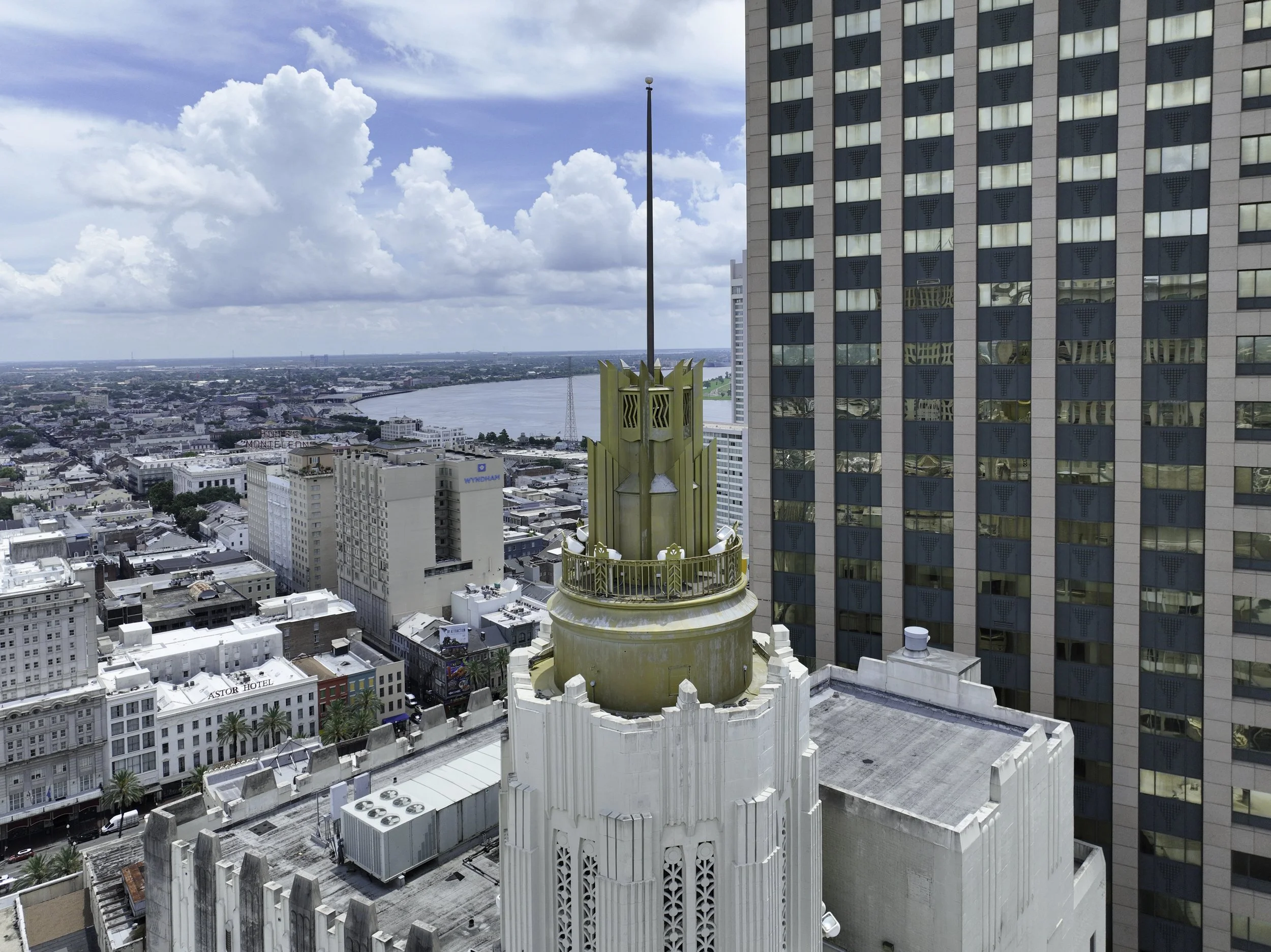 A cityscape view from a high rooftop, showing a large building with a golden decorative spire, surrounded by other buildings, a river in the background, and a partly cloudy sky.