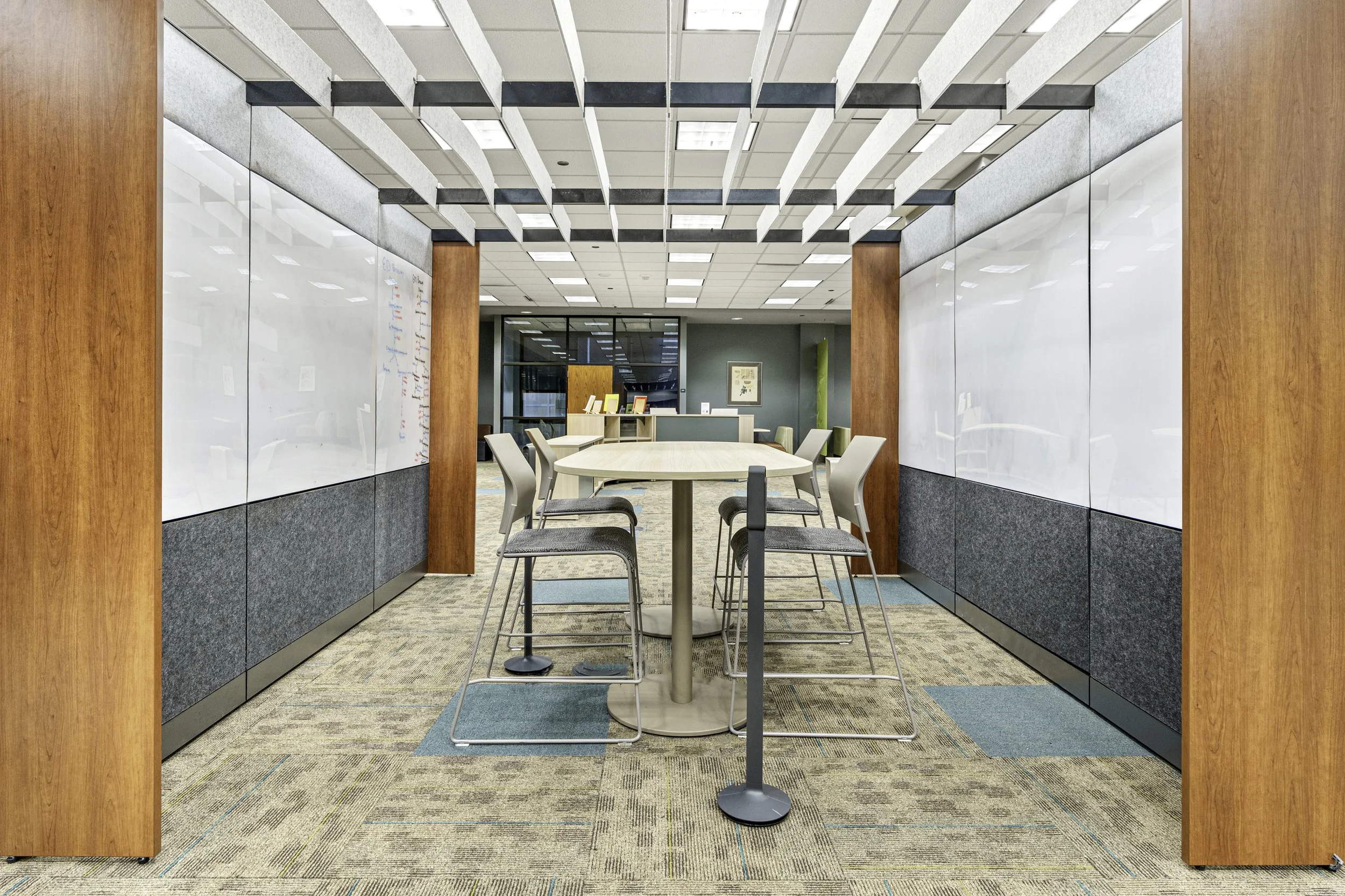 Meeting room with white round table, six chairs, glass walls, framed picture, and whiteboard on the left, with ceiling lights and carpeted floor.