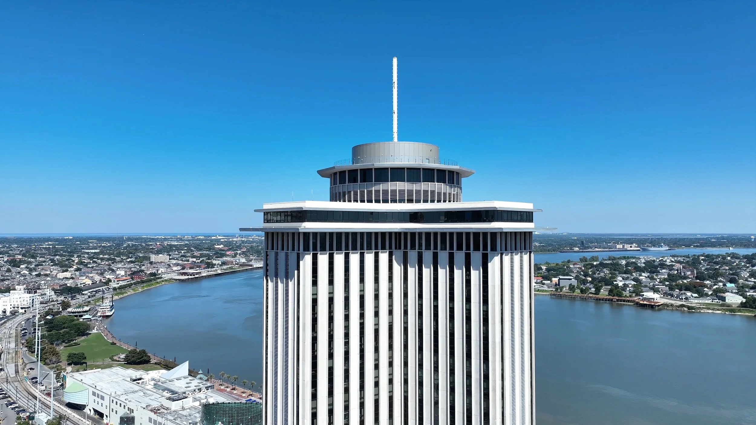 A tall modern skyscraper with a rounded top and an antenna on it, overlooking a river and a cityscape under a clear blue sky.