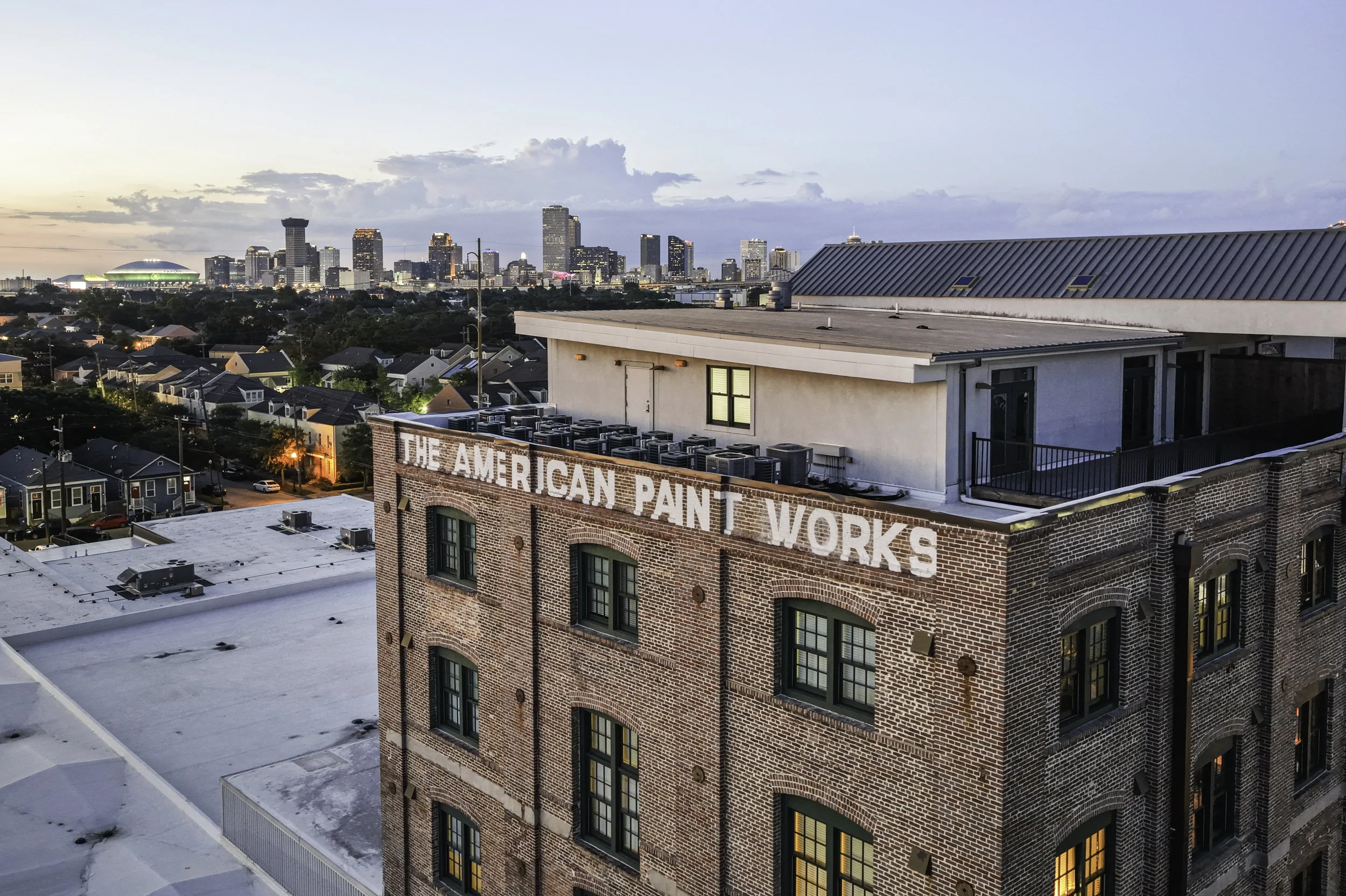 City skyline at dusk with high-rise buildings, stadium, and a rooftop view from a building with a sign that reads 'The American Paint Works'.