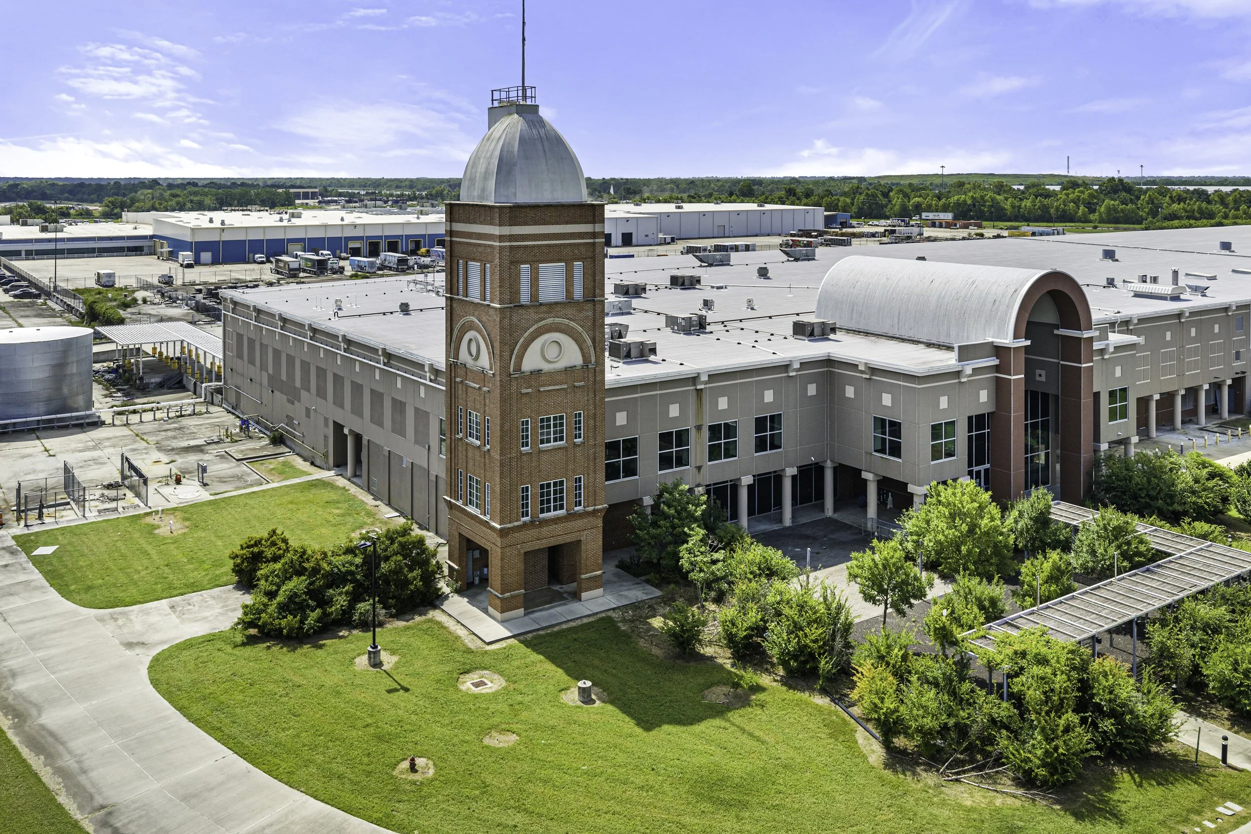 Aerial view of a large industrial or commercial building complex with a tall brick clock tower. The complex has multiple warehouses with white roofs, surrounded by parking lots, trucks, and greenery. In the background, there are trees and a blue sky with some clouds.