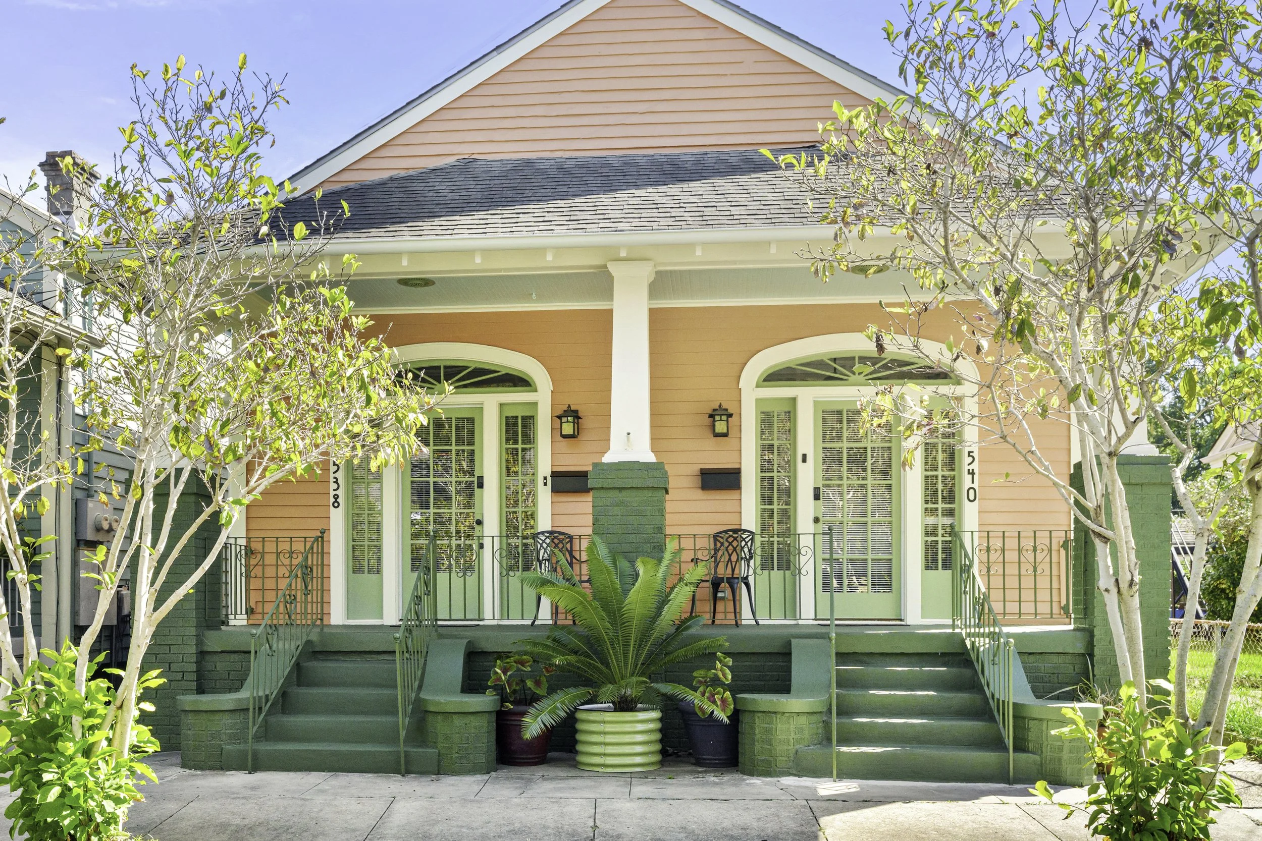 Front view of a two-story house with a peach-colored exterior, white trim, and green accents on the stairs, doors, and planters. The house has two front doors with glass panels, flanked by black lantern-style lights. Two trees with sparse leaves are 